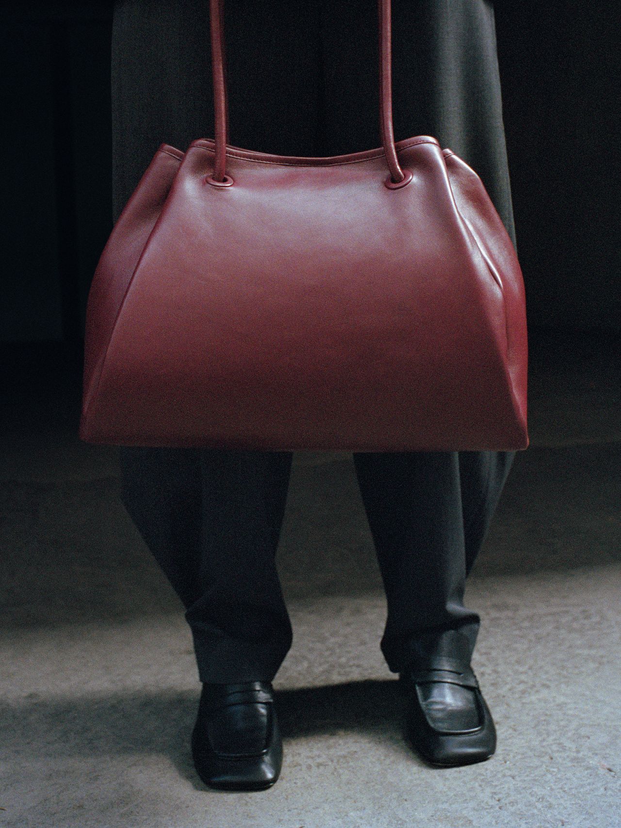 Person holding a large red leather handbag, wearing dark trousers and black shoes, standing on a concrete floor.