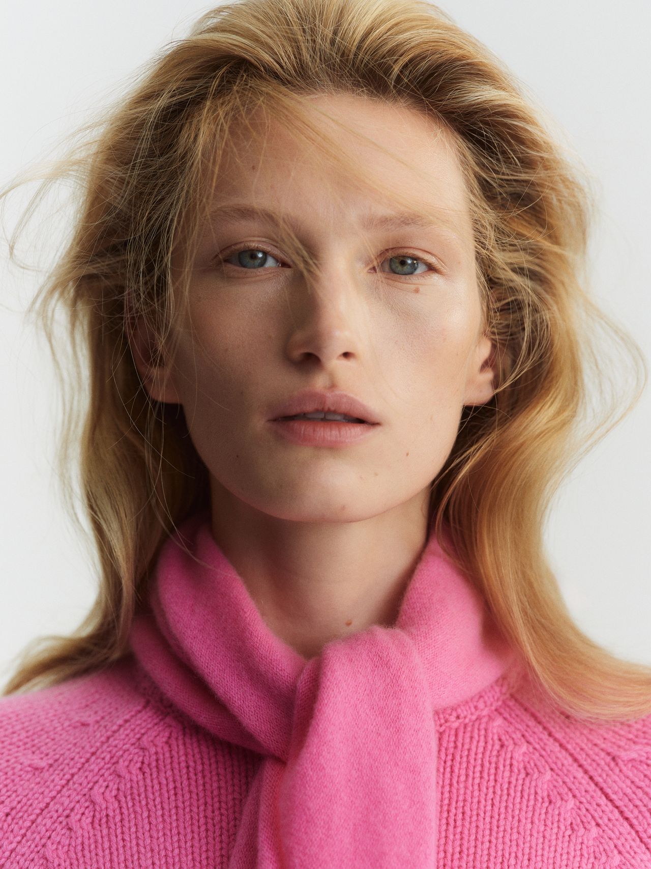 Person with long blonde hair wearing a pink sweater and scarf, looking directly at the camera against a plain background.