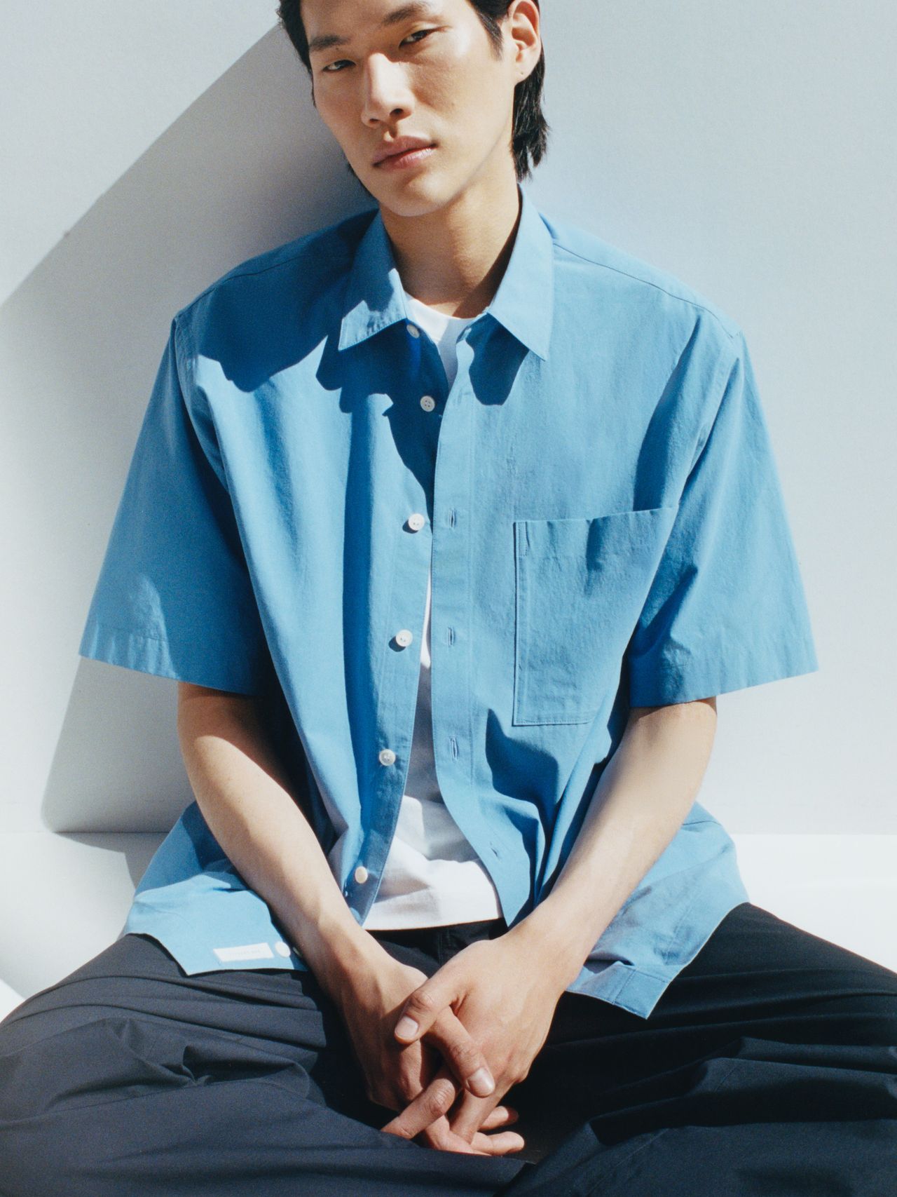 Person sitting against a white wall, wearing a light blue short-sleeve shirt over a white T-shirt, and dark pants, in a relaxed pose.