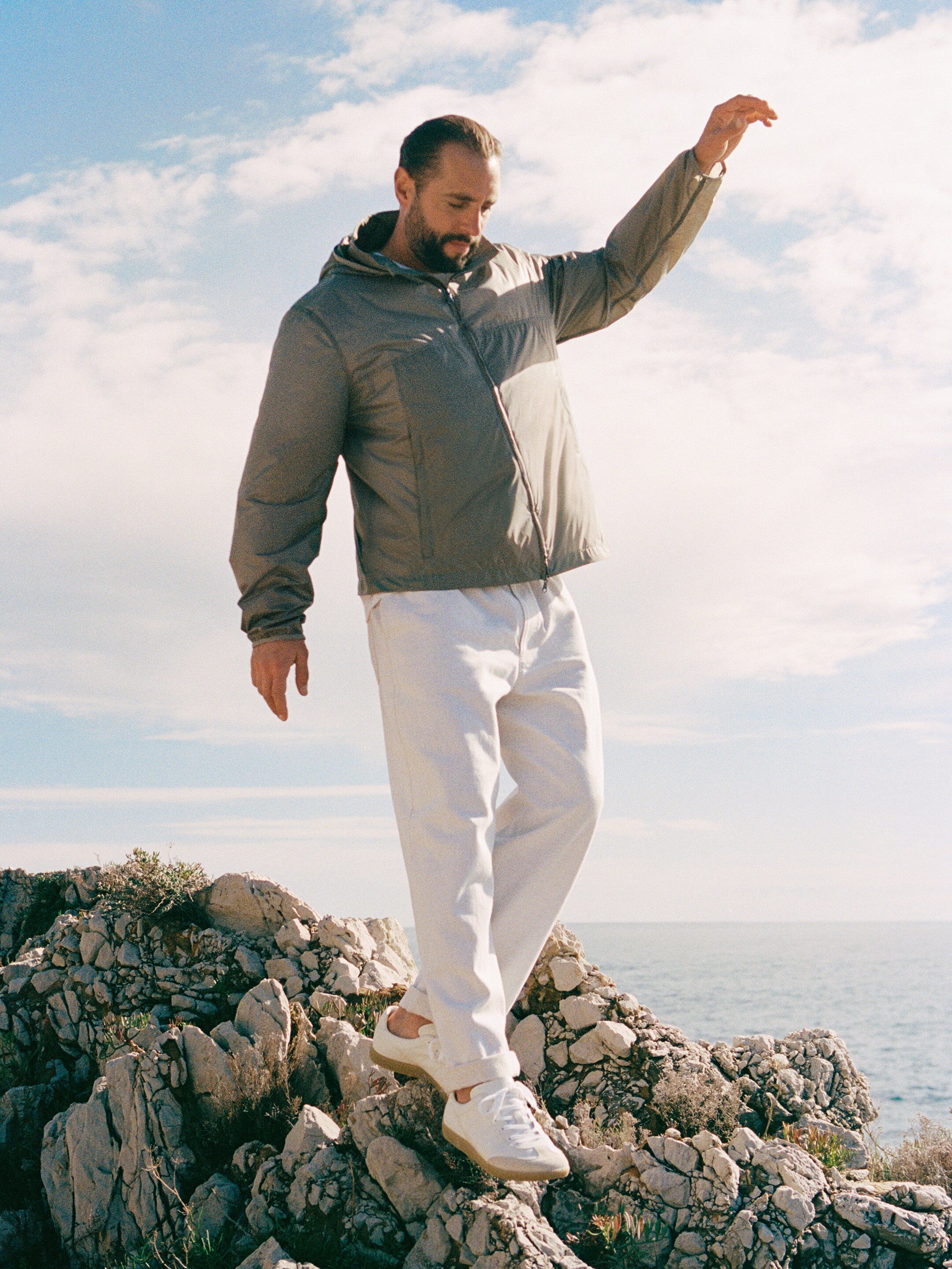 Man in a gray jacket and white pants balancing on rocky terrain by the sea under a partly cloudy sky.