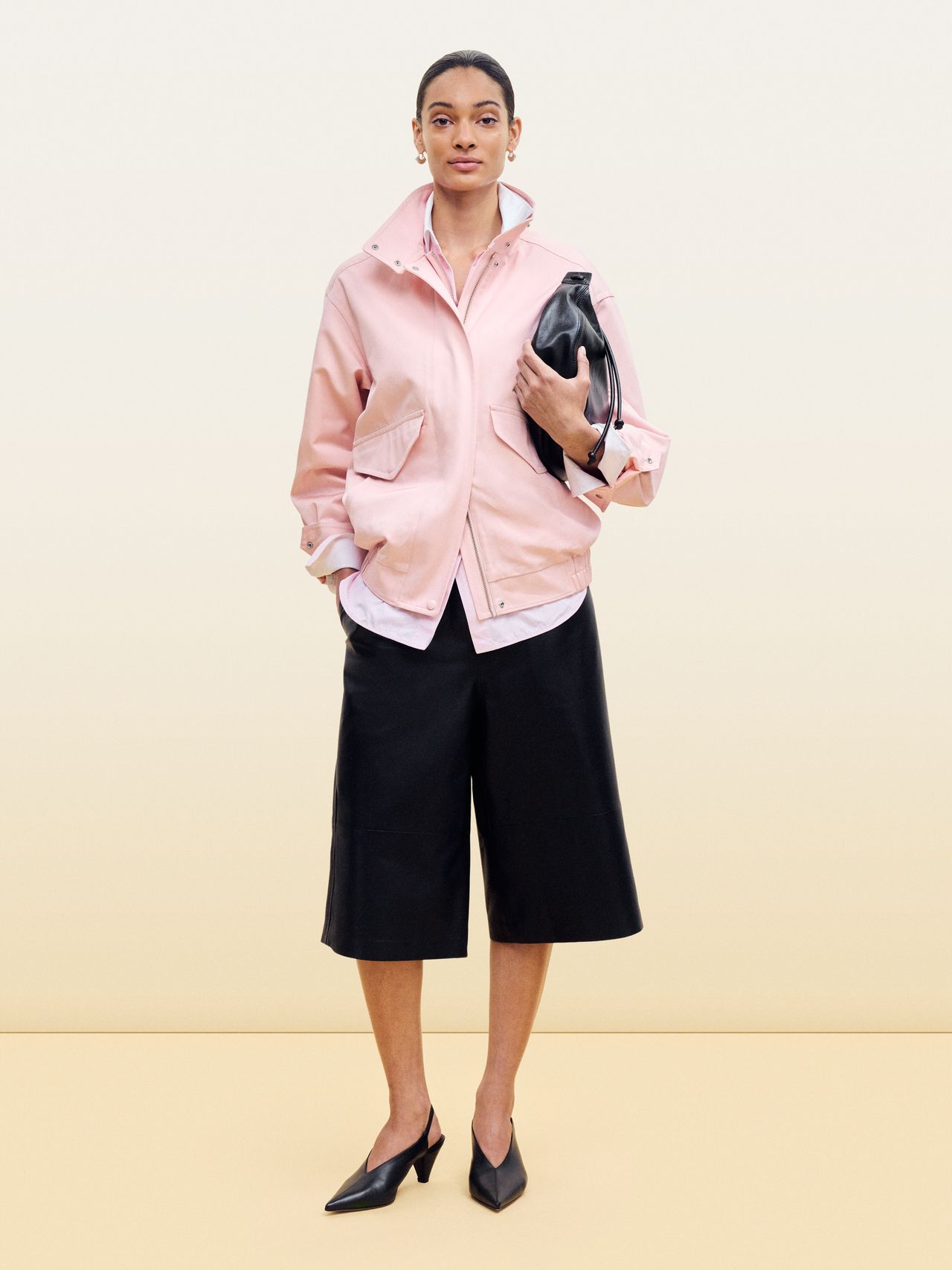 A person wearing a pink jacket, black culottes, and black heels, holding a black bag, stands against a light gradient background.