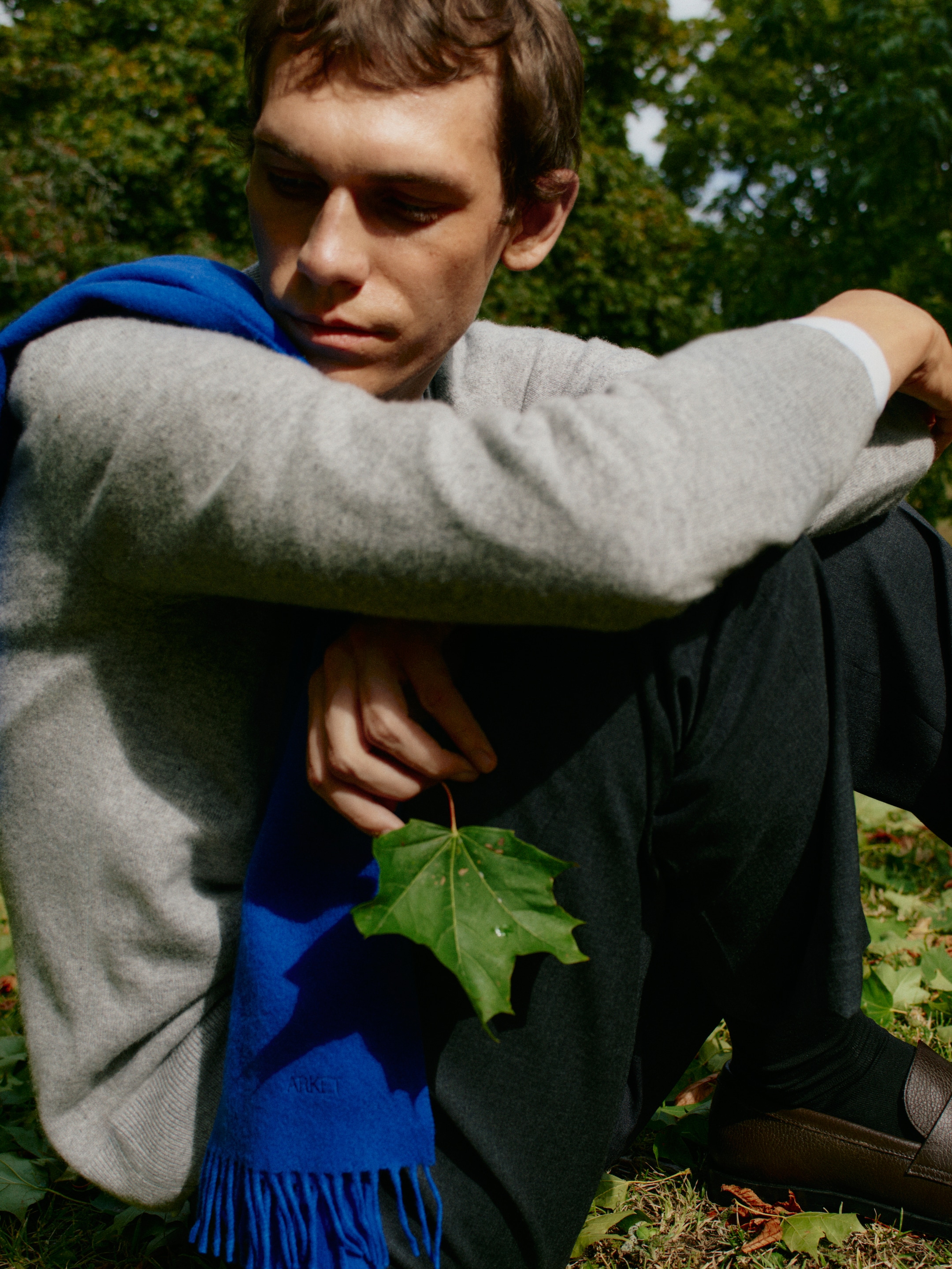 A person sitting on grass in a park, wearing a gray sweater and blue scarf, holding a leaf. The background is filled with green trees.