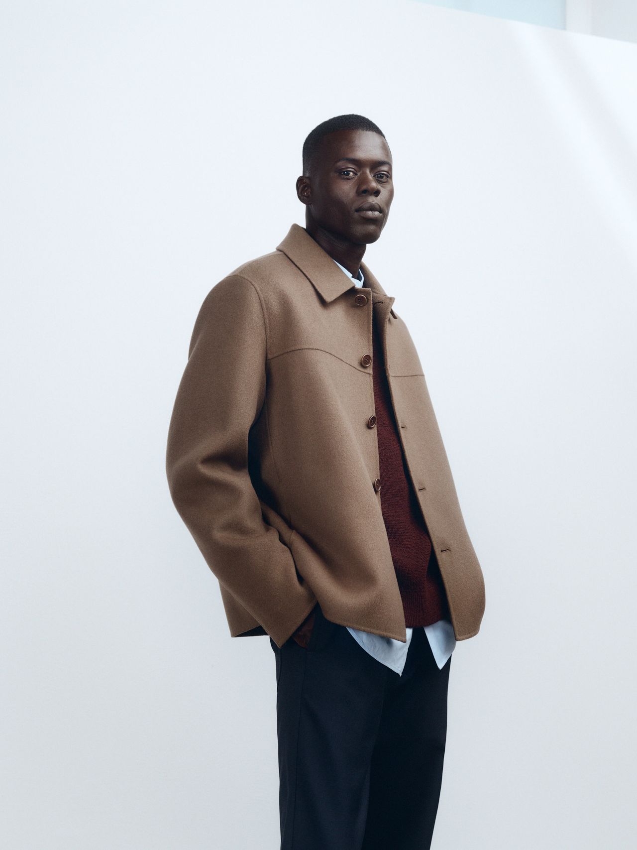 Person wearing a brown jacket over a red sweater and light blue shirt, standing against a plain white background.