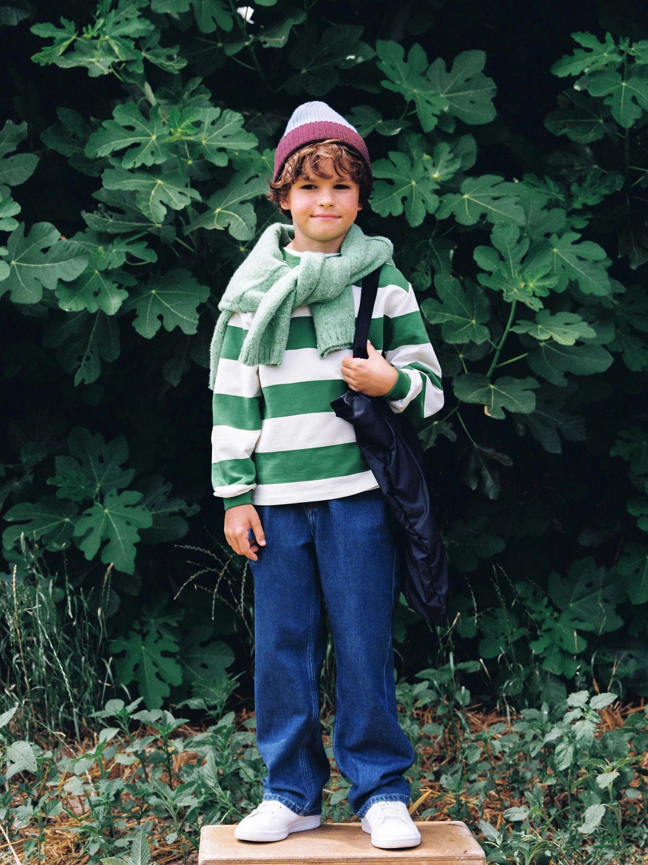 A child in a green and white striped sweater, jeans, and a beanie stands on a box against lush green foliage, holding a black bag.