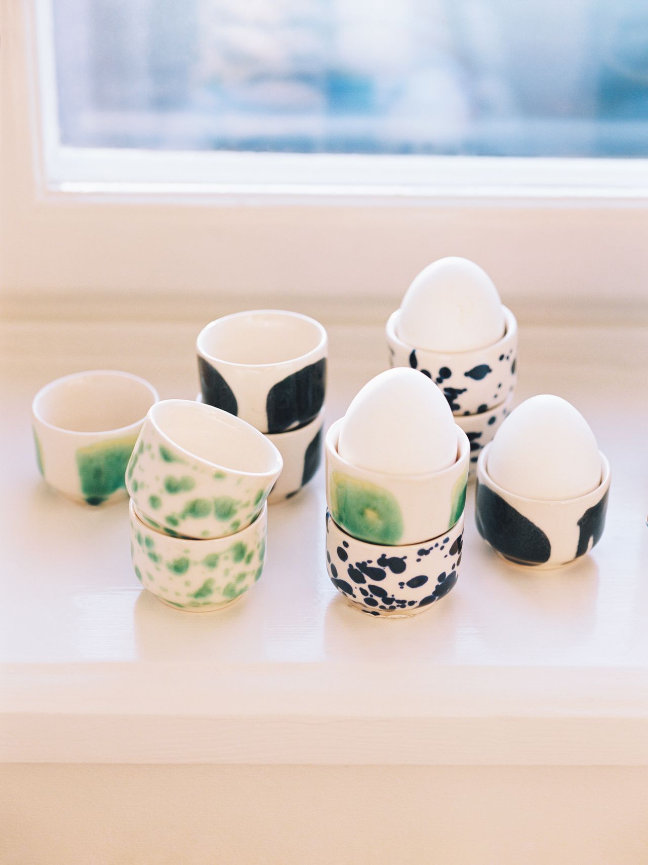 Ceramic egg cups with abstract black and green patterns, each holding a white egg, arranged on a white windowsill.