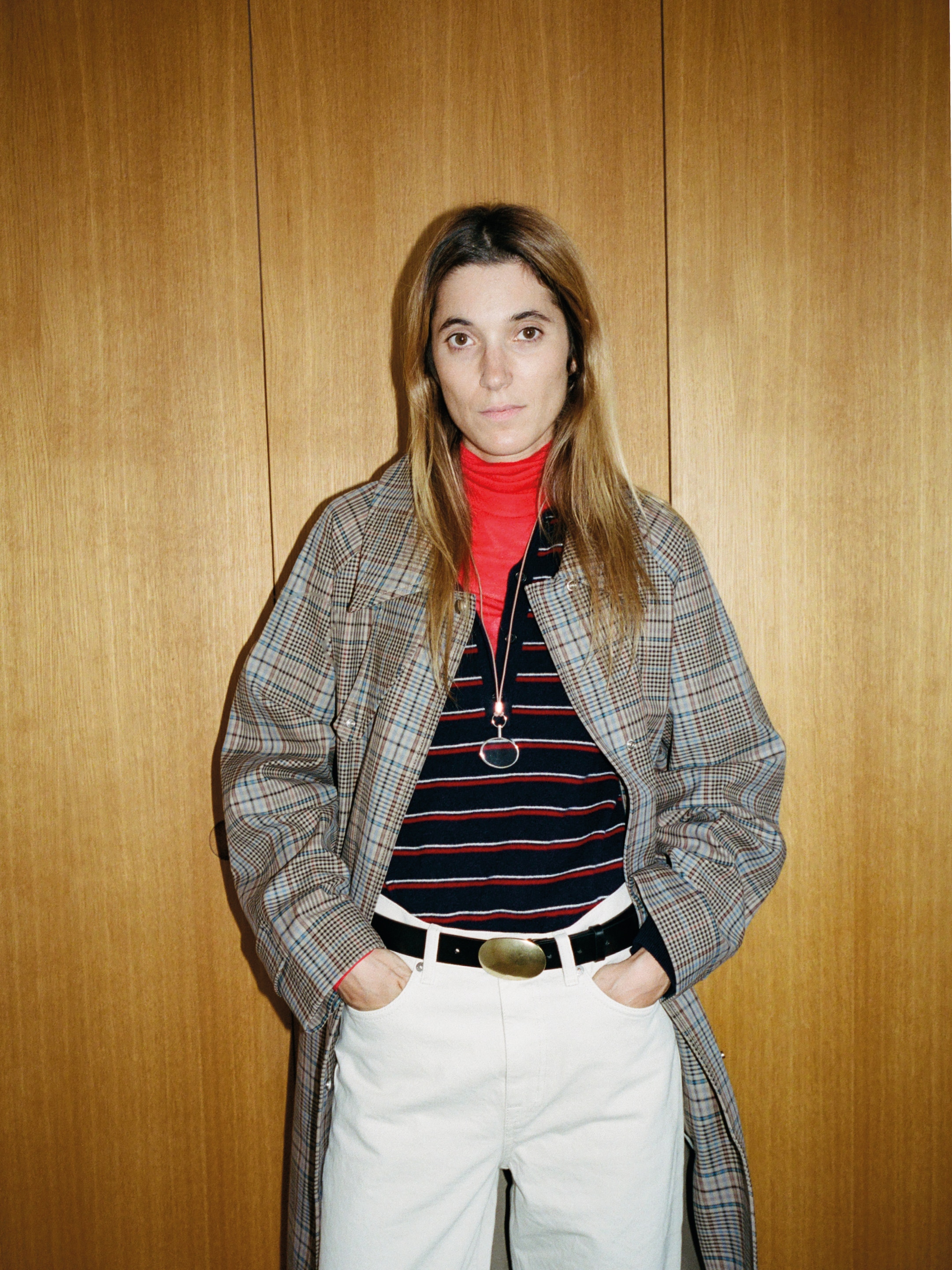 Woman with long hair in a plaid coat over a striped shirt, red turtleneck, and white pants, standing against a wooden background.