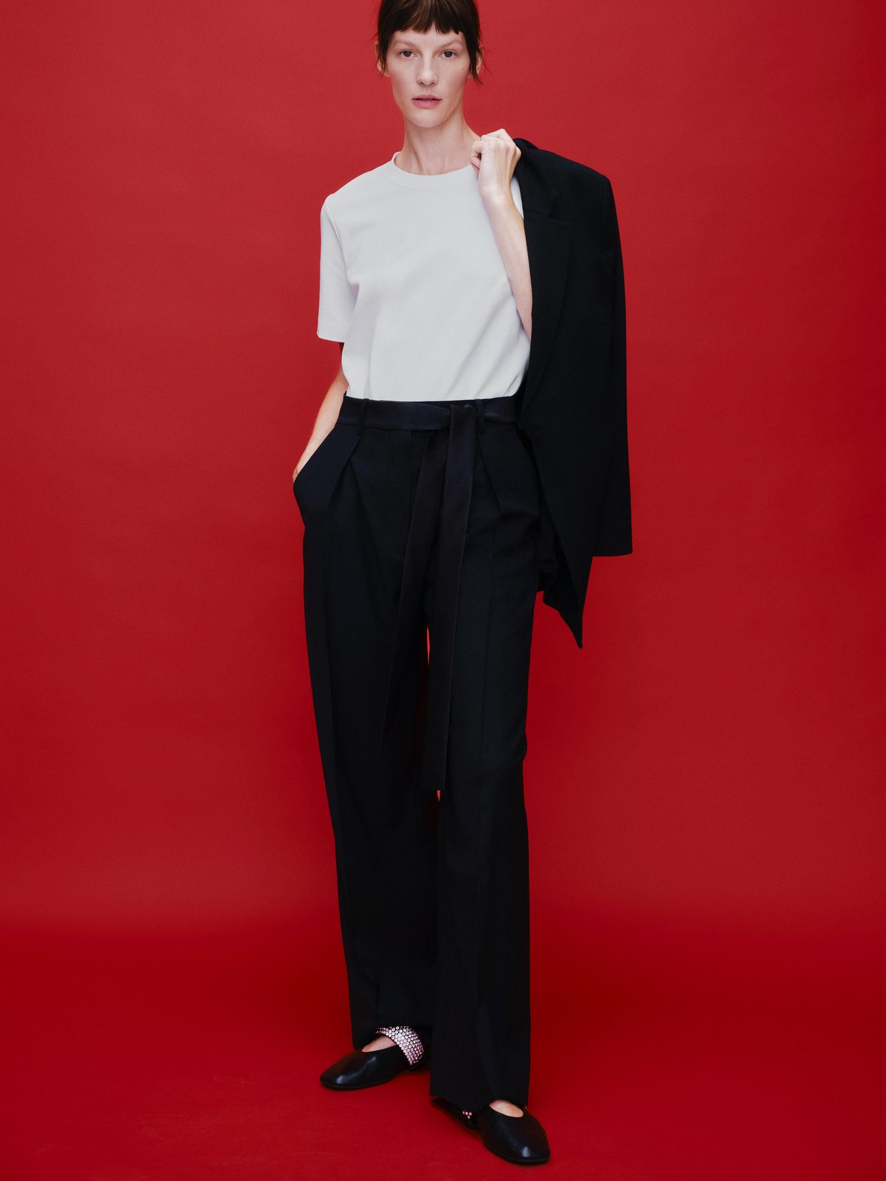 Person in a white shirt and black pants with a blazer over one shoulder, standing against a red background.