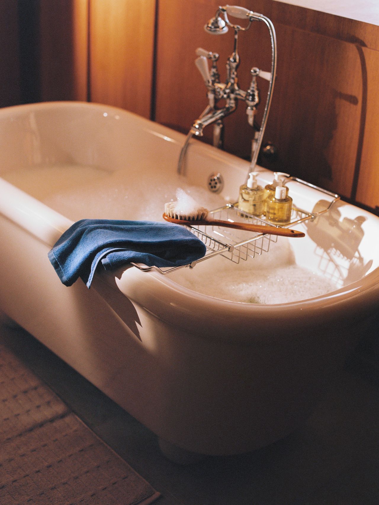 A vintage-style bathtub filled with bubbles, featuring a wooden tray holding a brush, soap, and bottles. A blue towel hangs over the side.