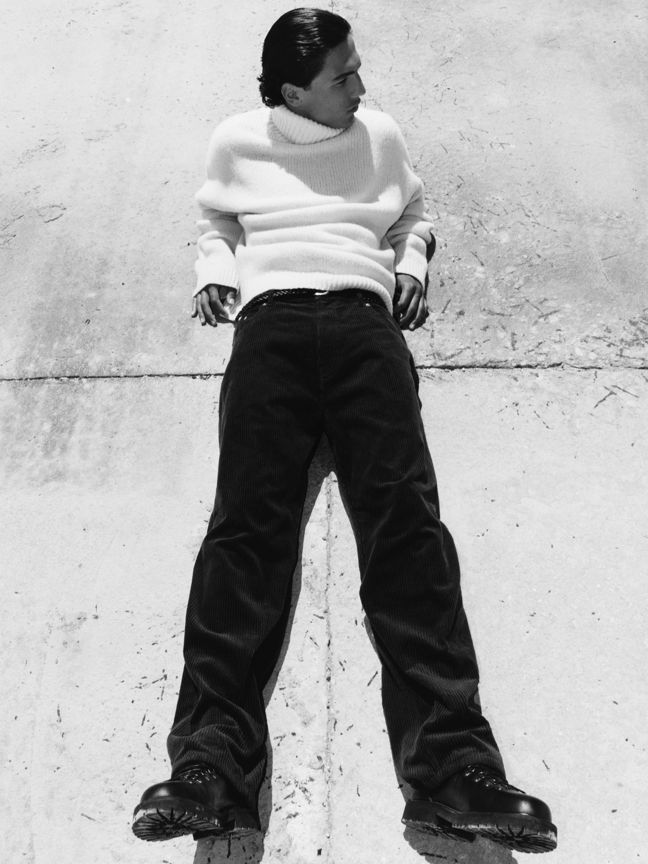 Person in a white turtleneck sweater and dark pants lying on a concrete surface, looking to the side. Black and white photo.
