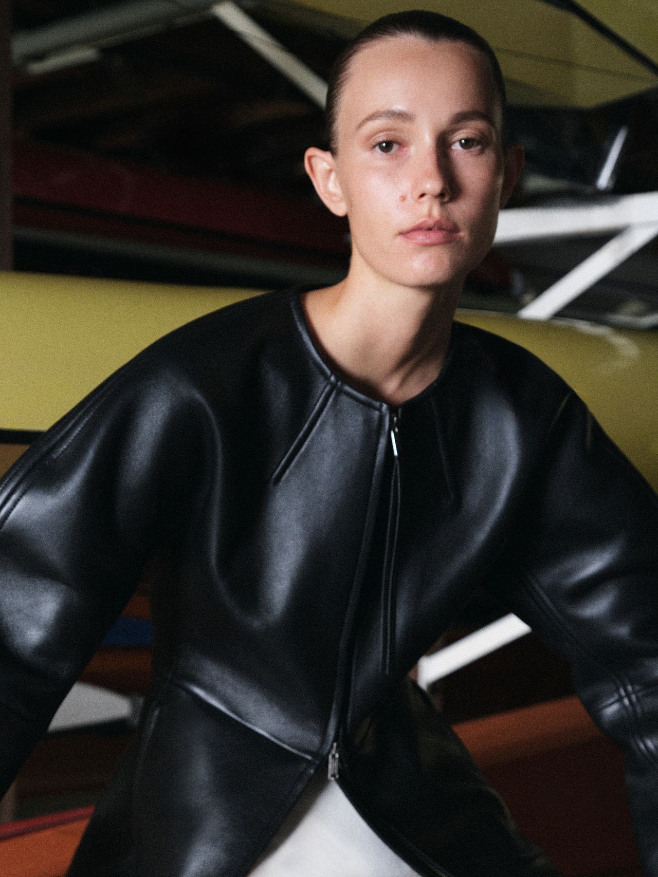 Person wearing a sleek black leather jacket, standing in an industrial setting with a focused expression.