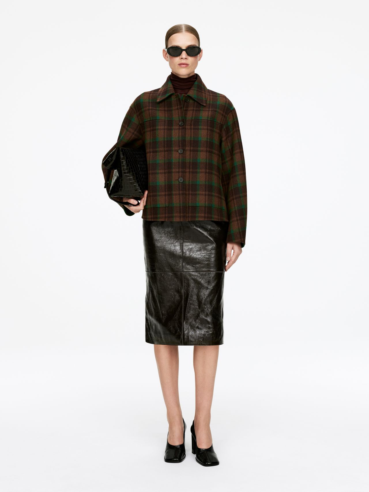 Person wearing sunglasses, a plaid jacket, and a black leather skirt, holding a textured black handbag, standing against a plain background.