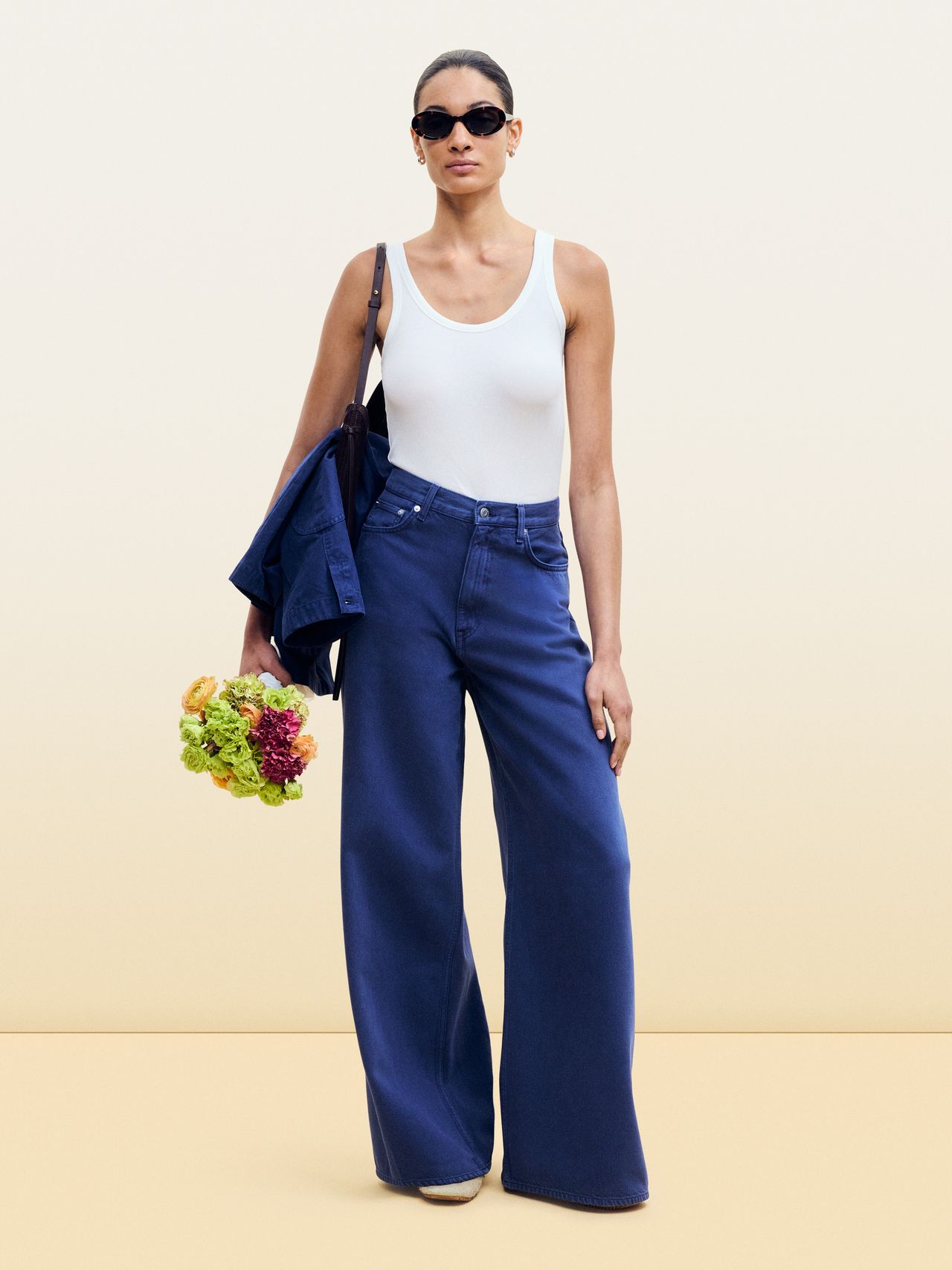 Woman in sunglasses wearing a white tank top and wide-leg blue jeans, holding a bouquet and a jacket against a plain background.