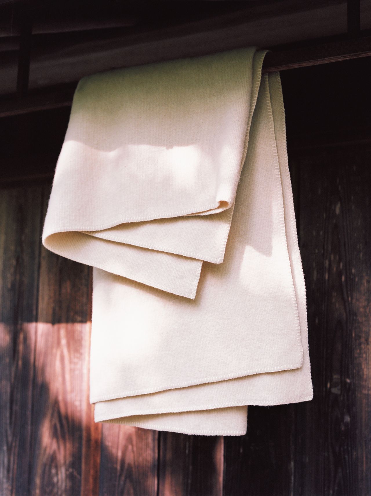 A beige blanket hangs over a wooden railing, with soft sunlight casting shadows on its surface. The background features rustic wooden panels.