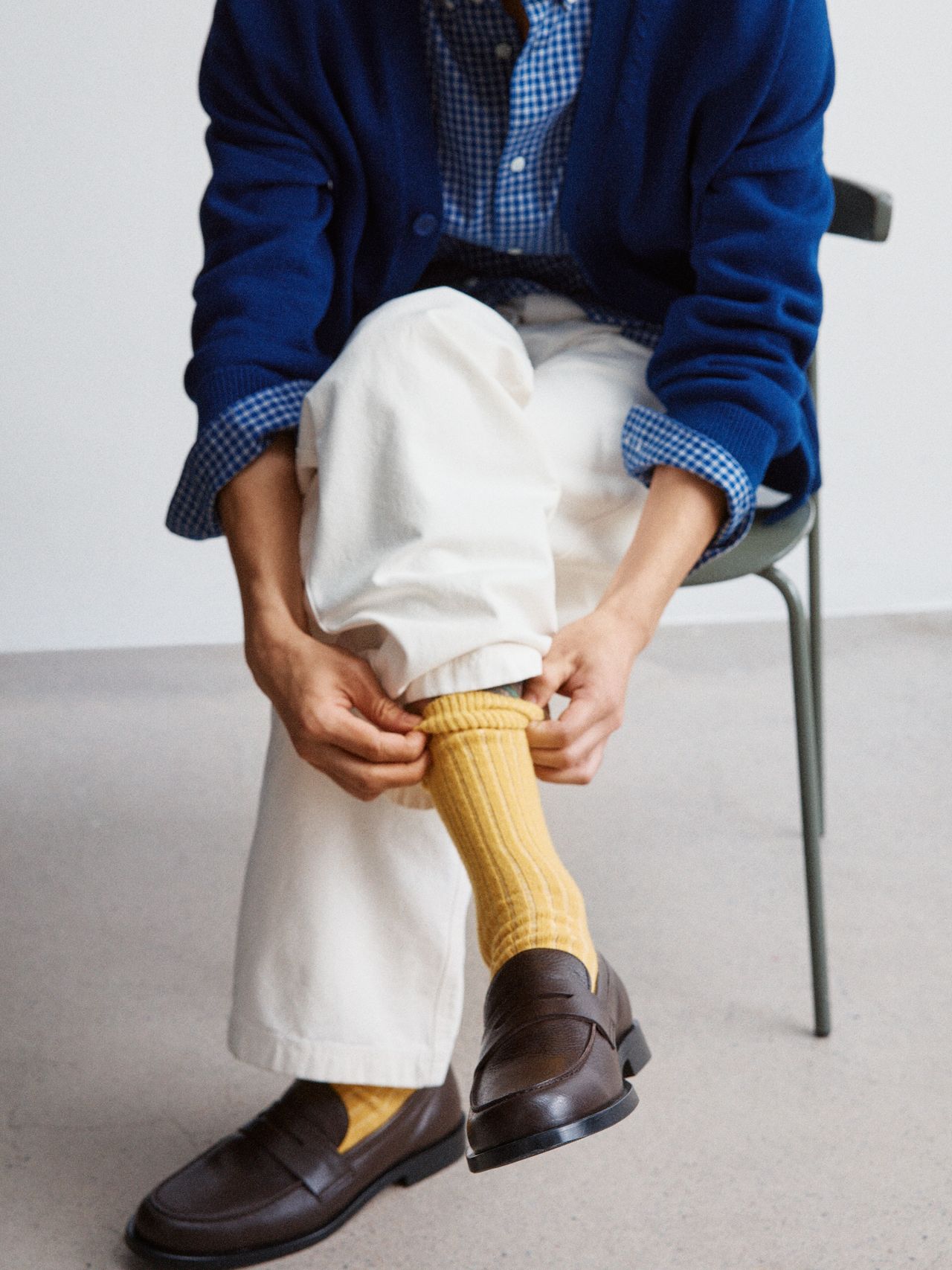 Person seated, adjusting yellow socks. They wear a blue cardigan over a checkered shirt, cream pants, and brown loafers.