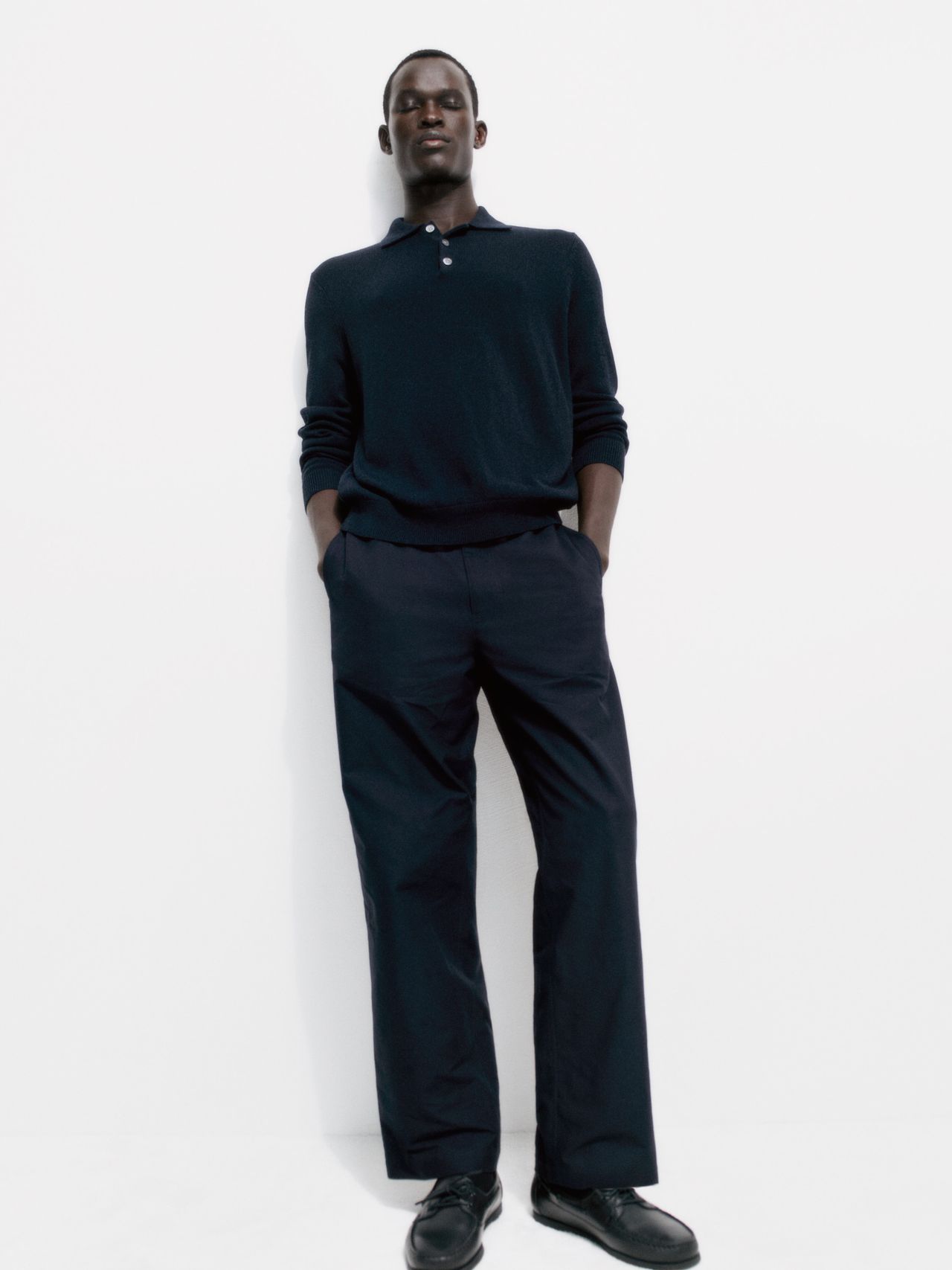 Person in navy attire stands against a plain white background, hands in pockets, exuding a relaxed and stylish demeanor.