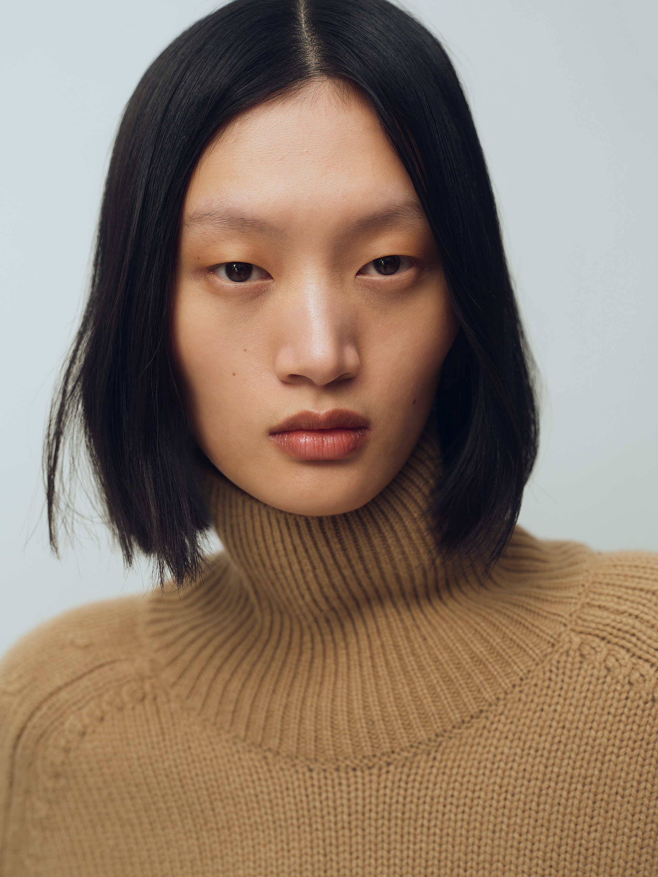 A person with straight black hair wears a camel-colored turtleneck sweater against a neutral background.
