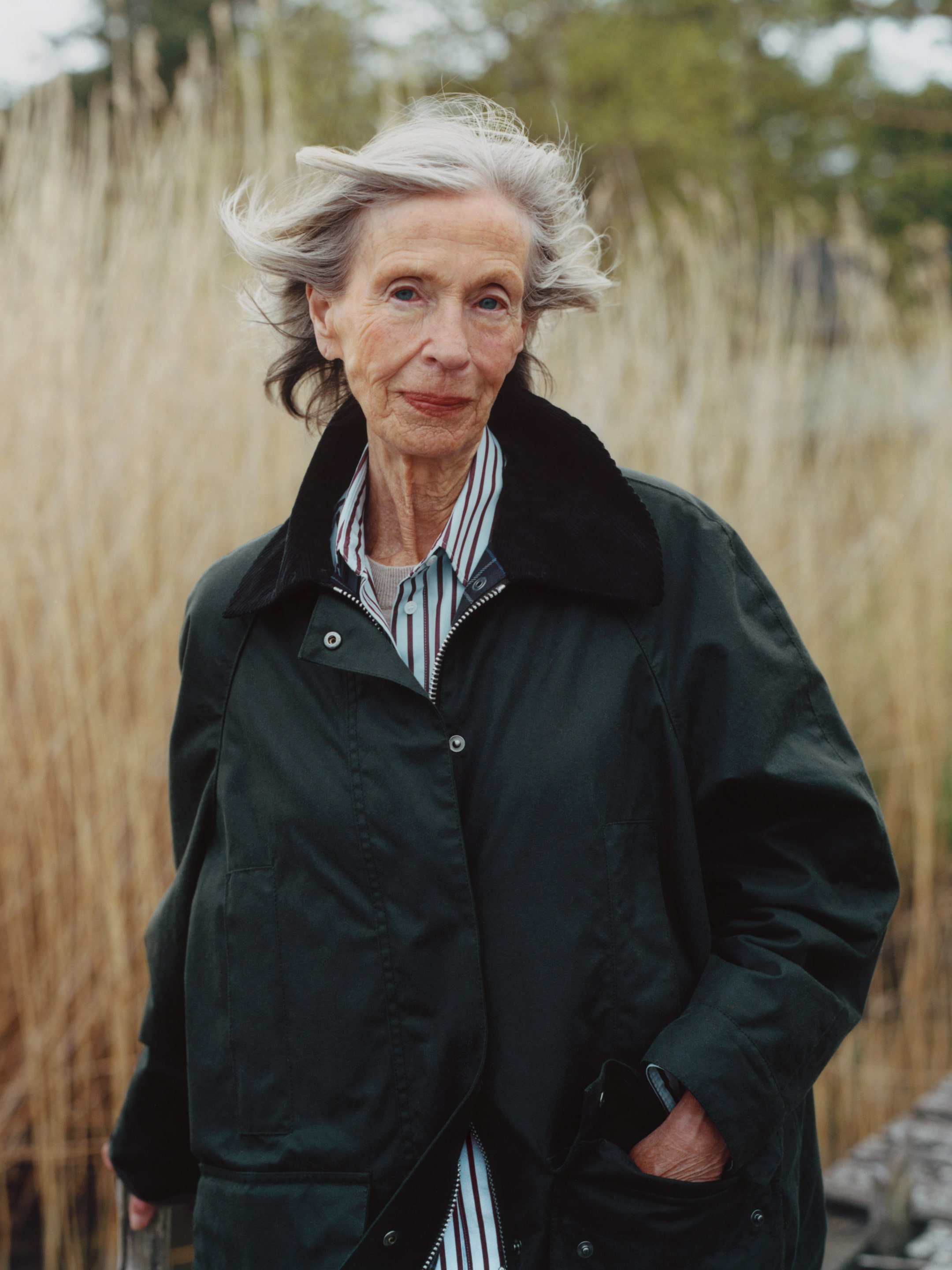Elderly woman with gray hair wearing a dark coat stands outdoors, wind blowing her hair, with tall grasses in the background.