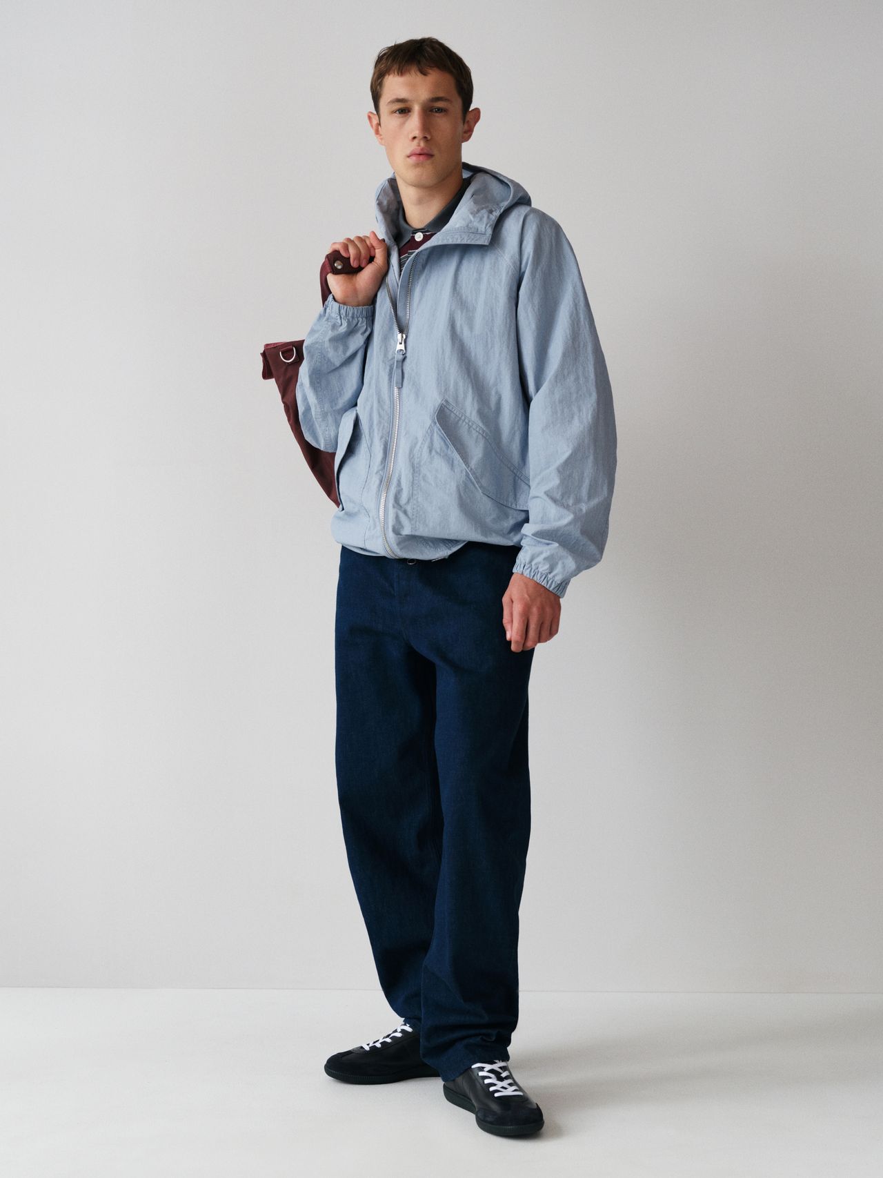 Person wearing a light blue hooded jacket, dark pants, and black sneakers, holding a maroon bag against a plain white background.