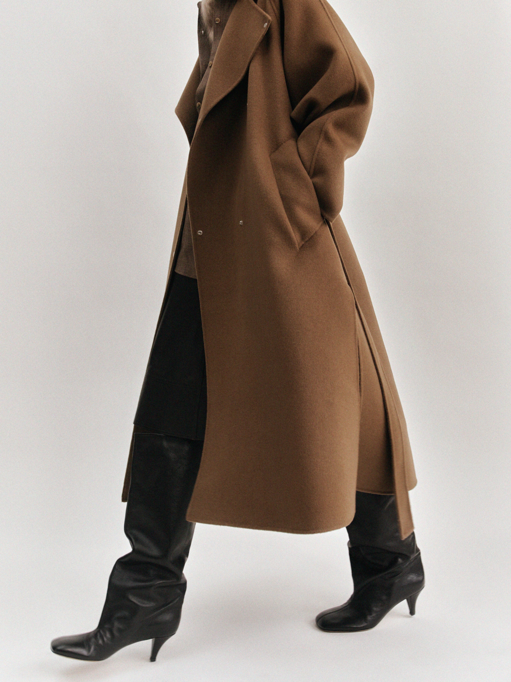 Person in a brown overcoat and black knee-high leather boots, walking against a light background.