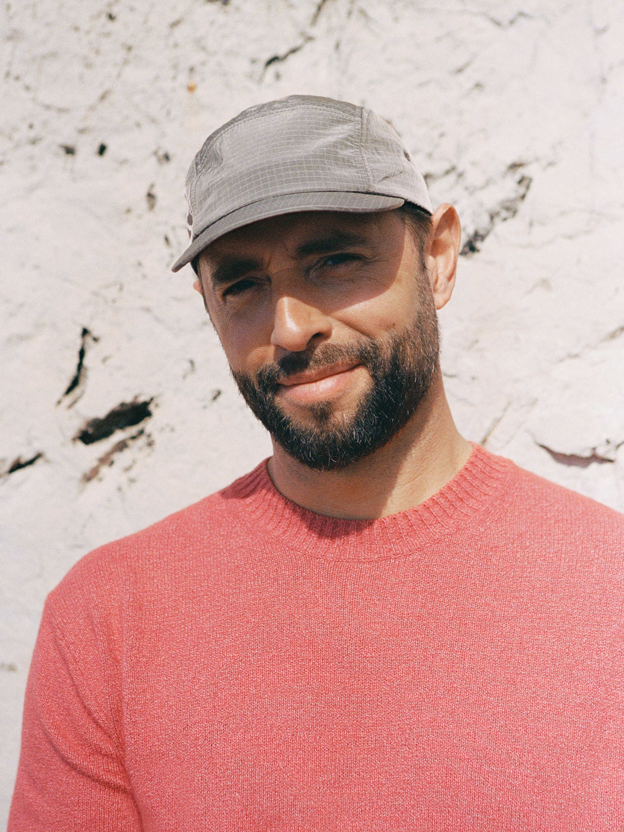 Man with a beard wearing a gray cap and pink sweater, standing against a textured white wall, smiling slightly.