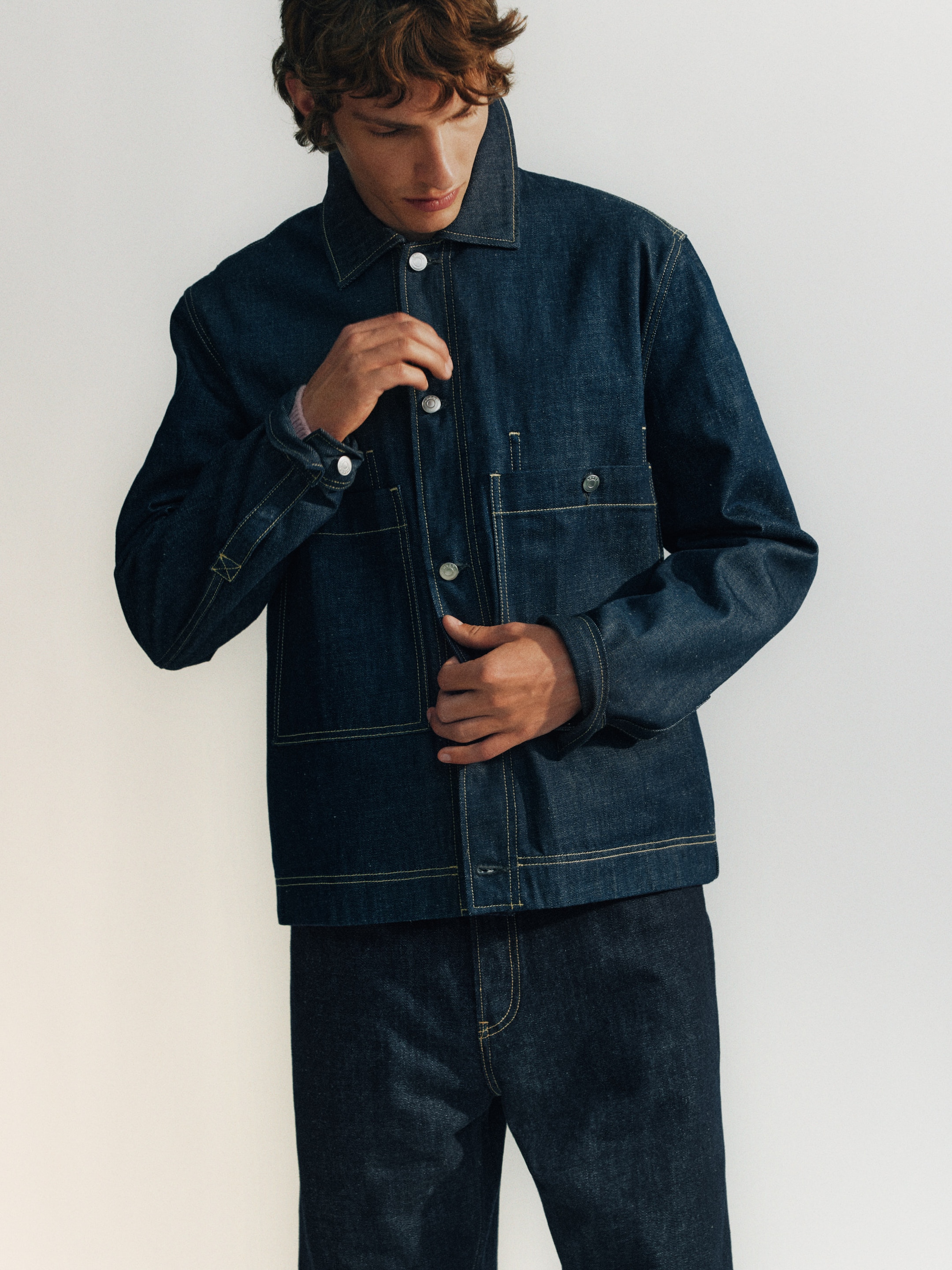 Person wearing dark denim jacket and jeans, buttoning the jacket. They have curly brown hair and are looking down slightly. Simple background.