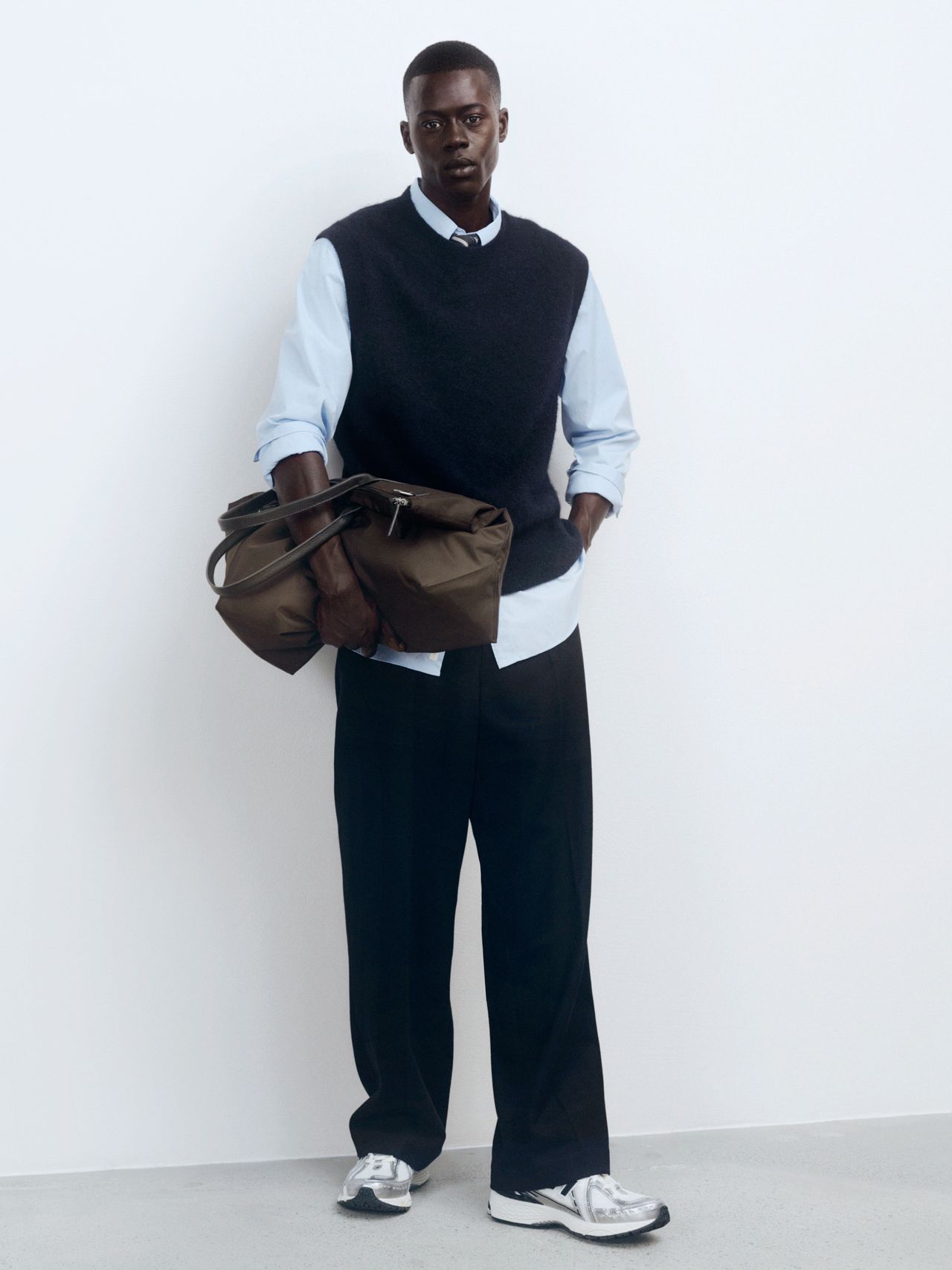 Man in a blue shirt, dark sweater vest, and black trousers holds a brown bag, standing against a white wall.