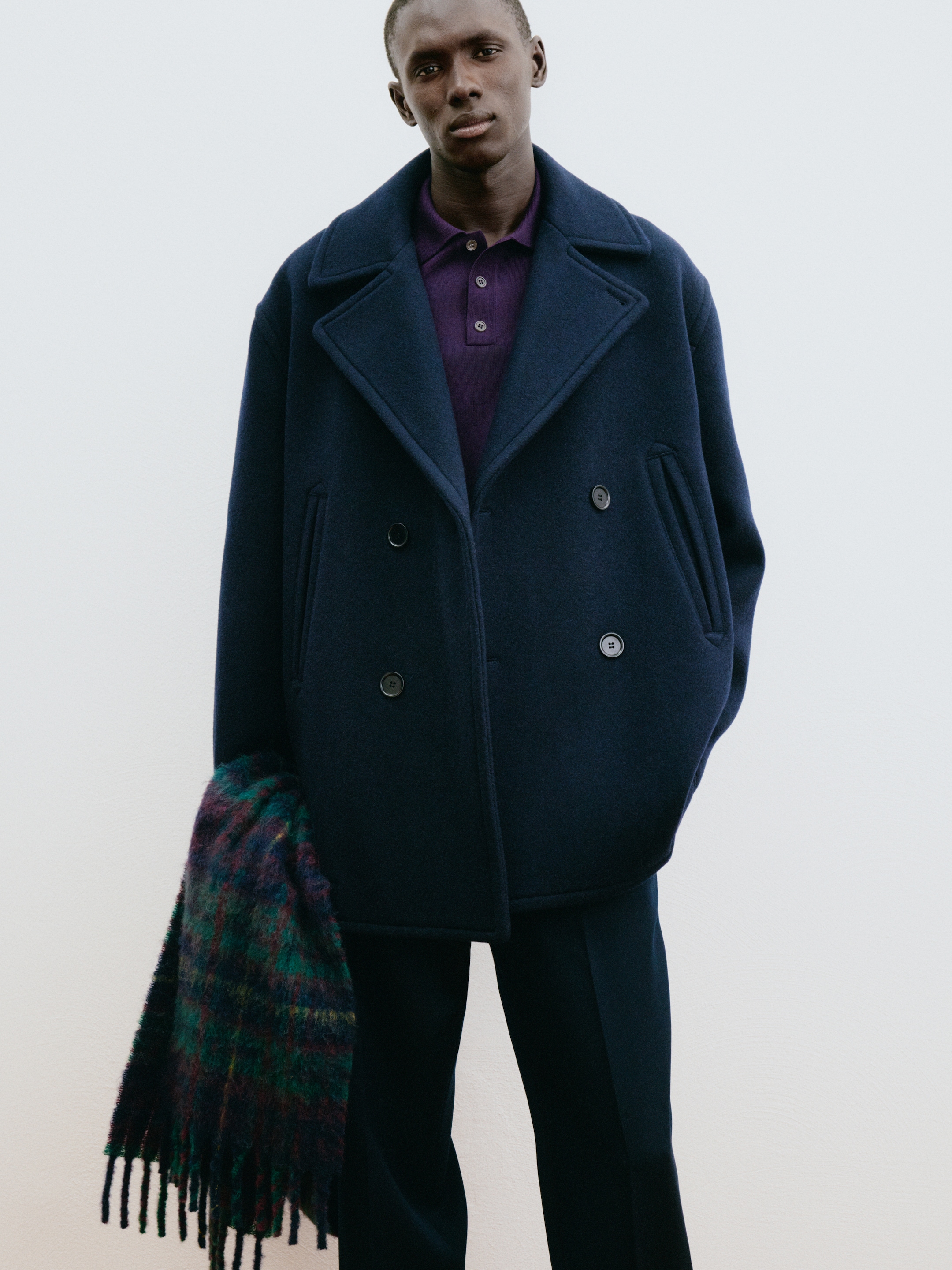 Person wearing a navy pea coat and purple shirt, holding a multicolored plaid scarf against a plain background.
