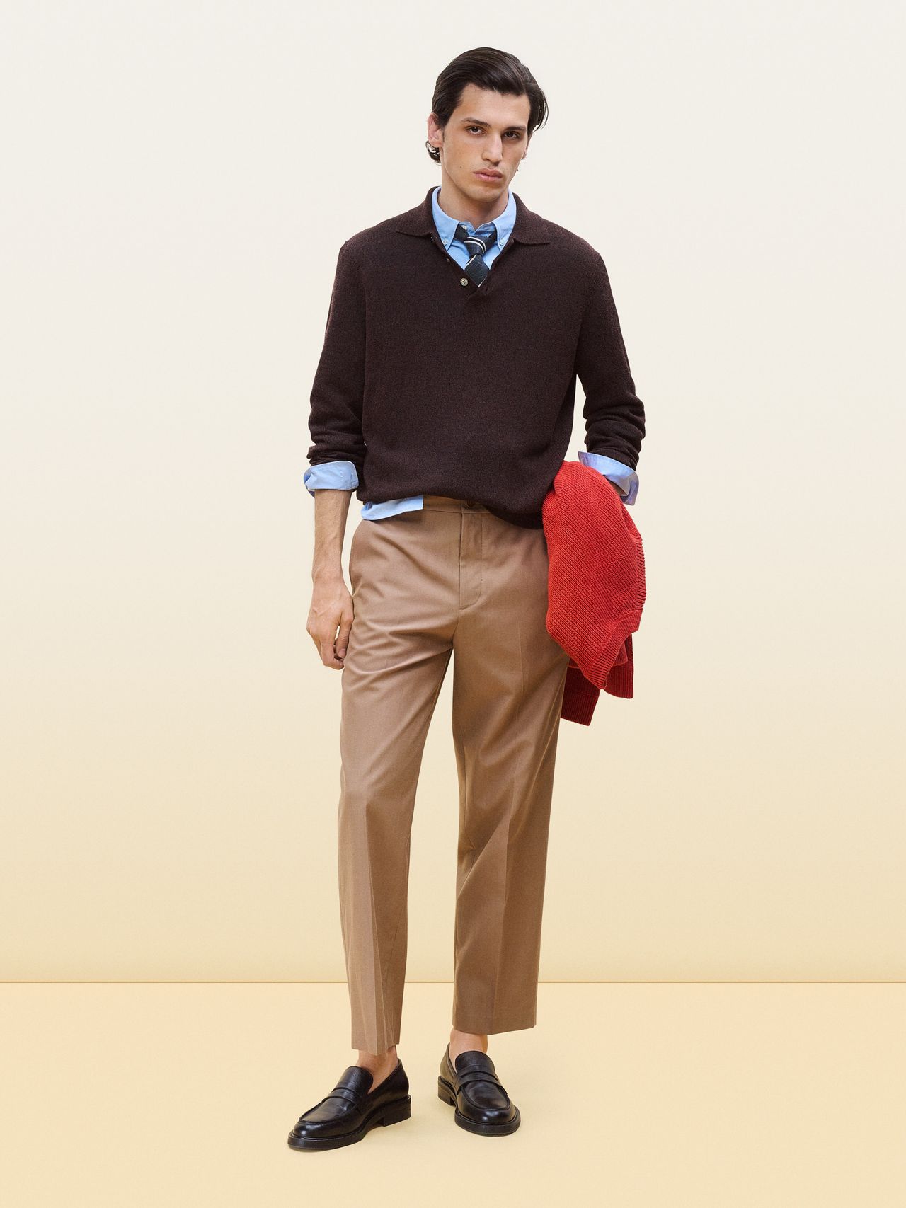 Man in a dark sweater over a blue shirt and tie, holding a red jacket. He wears beige trousers and black loafers. Neutral backdrop.