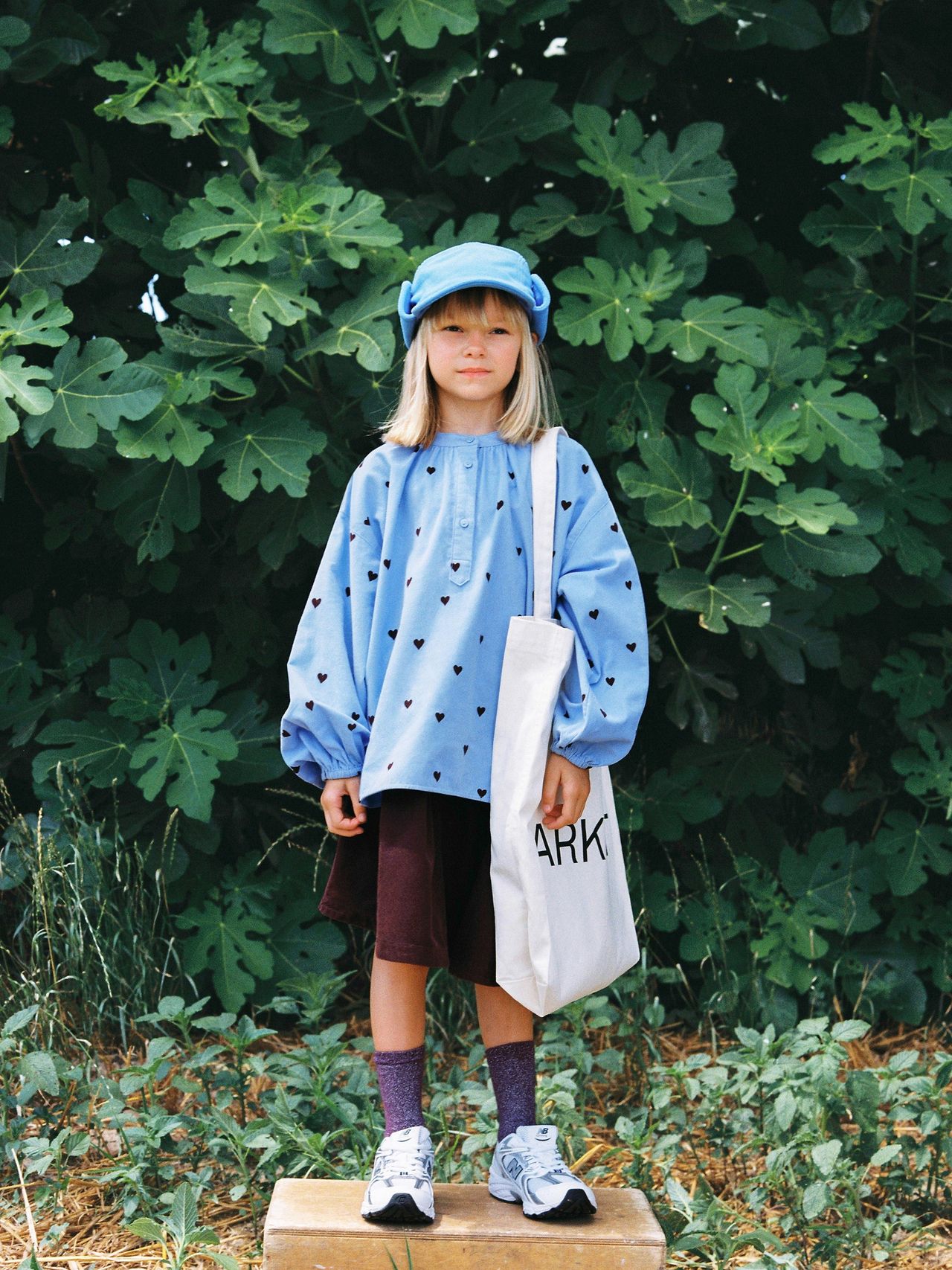 A young child in a blue outfit and cap stands on a wooden block, holding a white tote bag, with lush green foliage in the background.