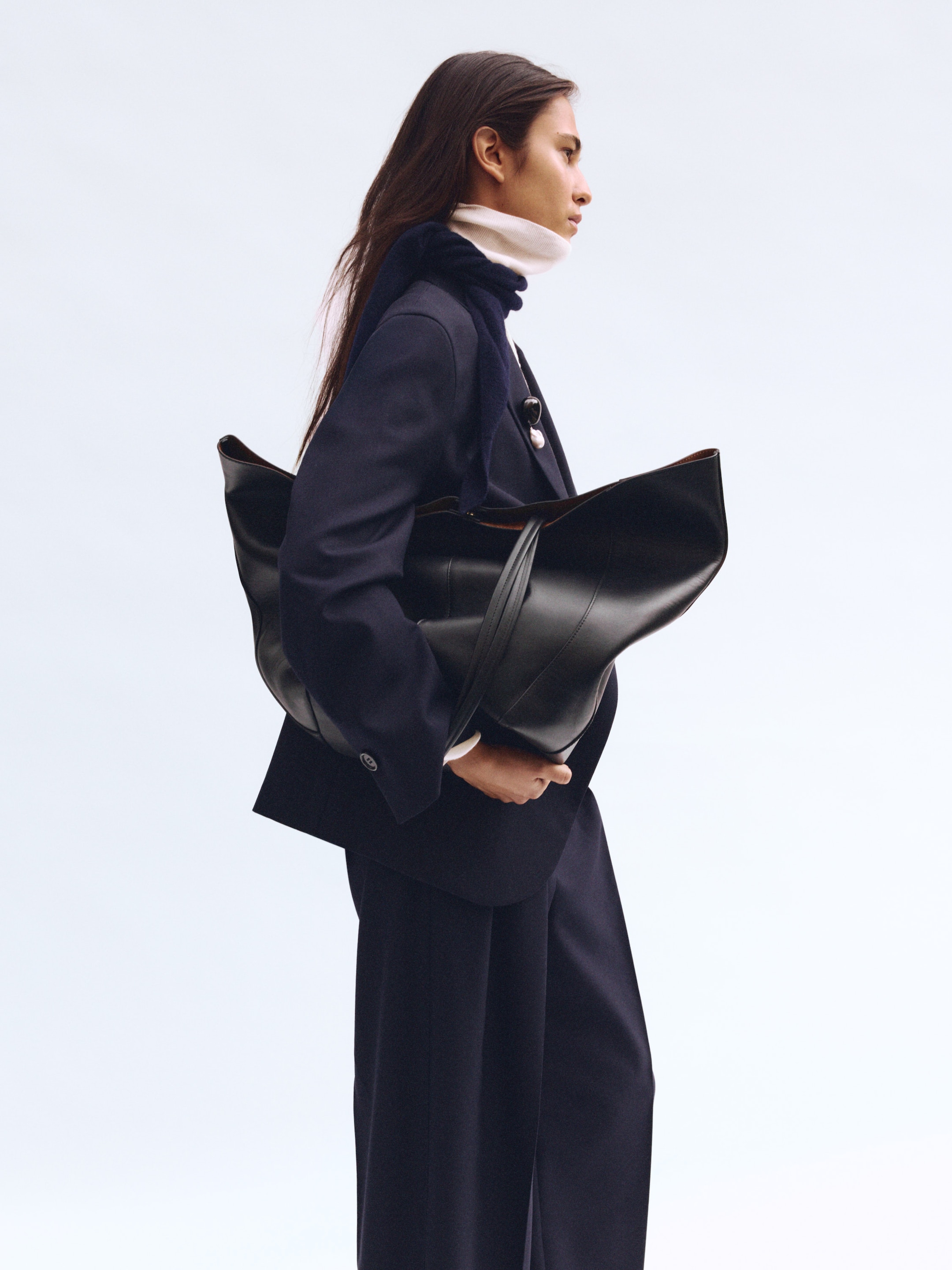 A woman in a navy outfit and white turtleneck holds a large black bag, standing against a plain white background.
