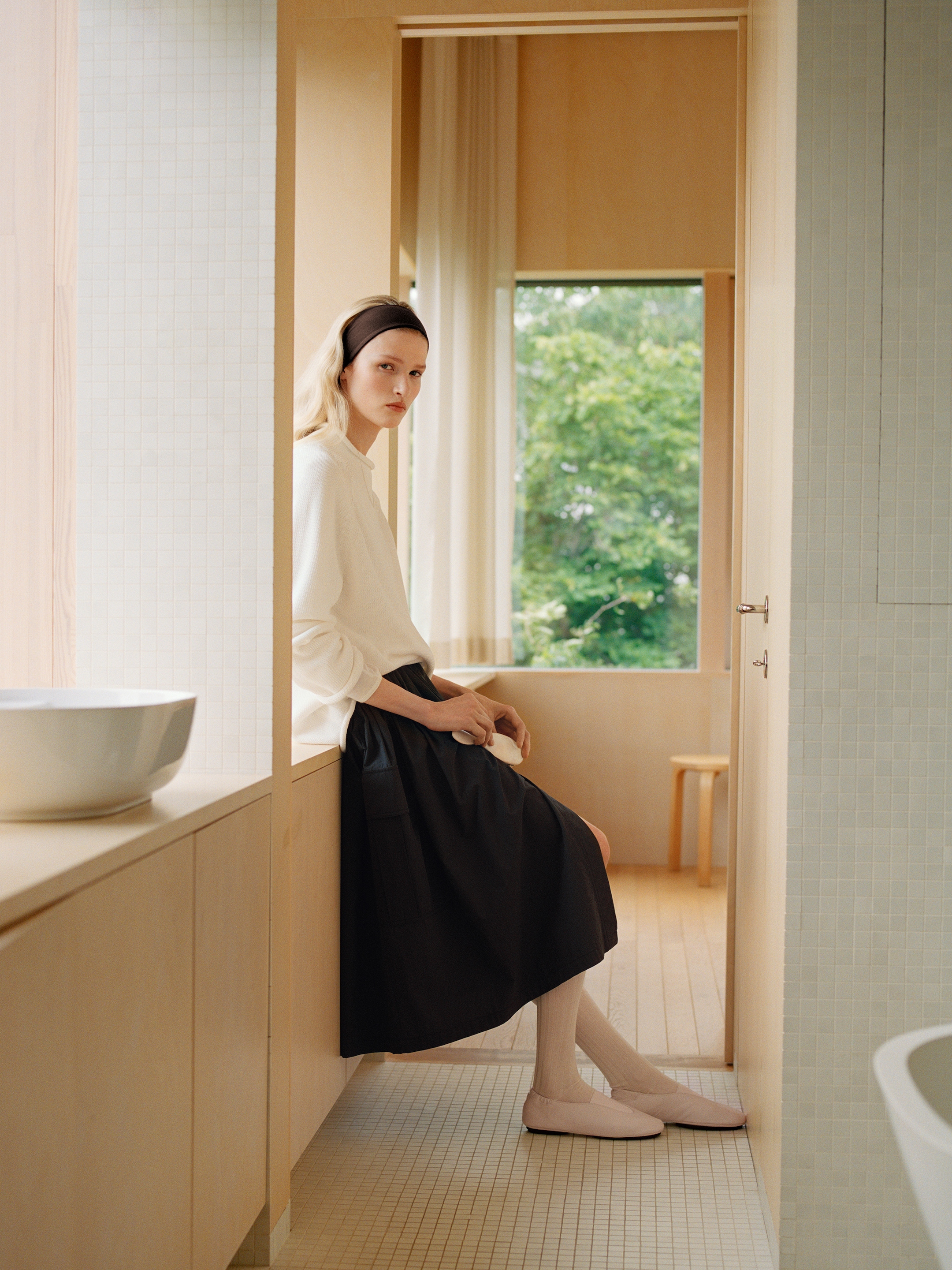 A woman sits in a minimalist room with light wooden walls and tile floors, wearing a white top, dark skirt, and headband, near a window overlooking greenery.