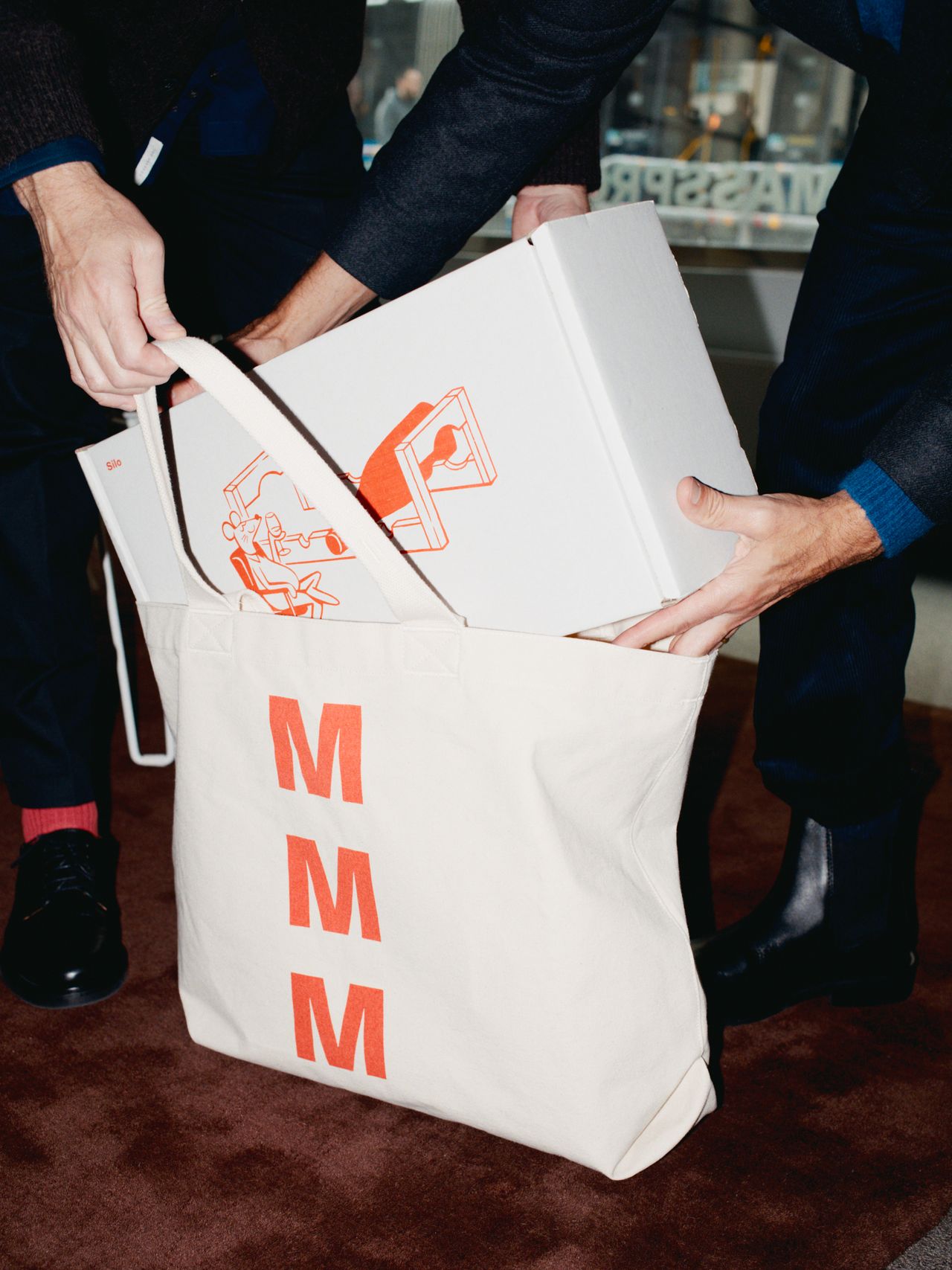 Two people placing a white box with orange graphics into a cream tote bag with "MMM" in red letters, on a brown surface.