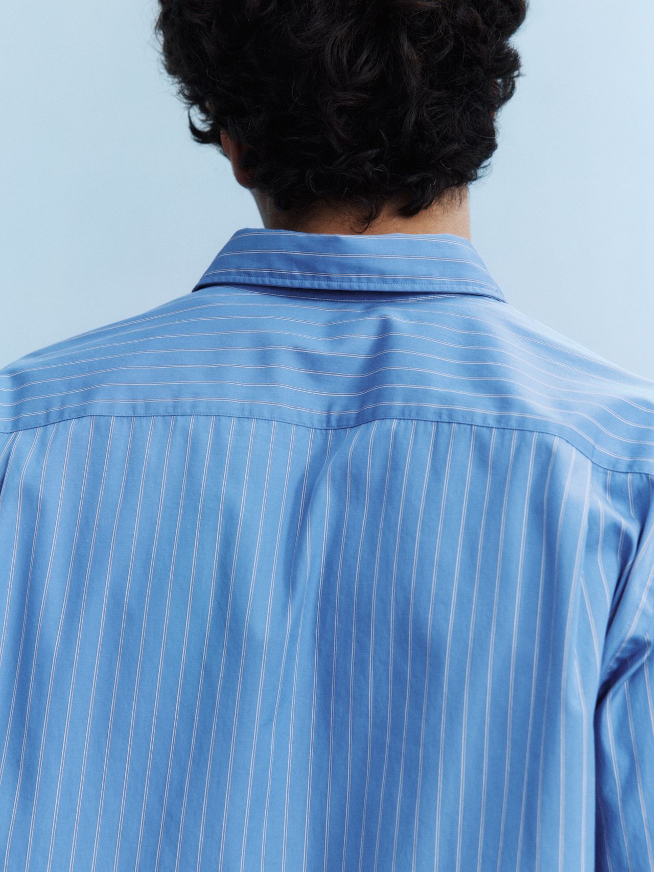 Person with curly hair wearing a blue pinstripe shirt, seen from behind against a light blue background.