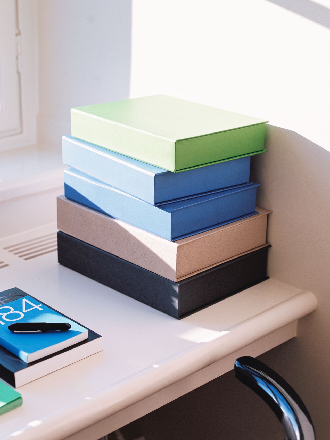 A stack of colorful books on a white desk, with a pen and another book nearby. Sunlight casts shadows across the surface.