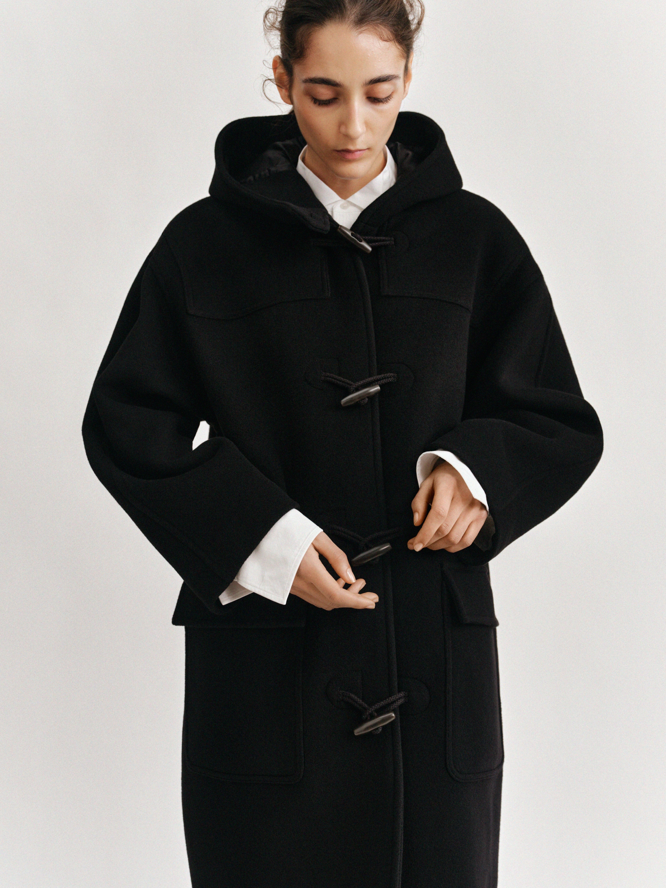 A person wearing a black duffle coat with a hood and toggles stands against a plain background, looking down.
