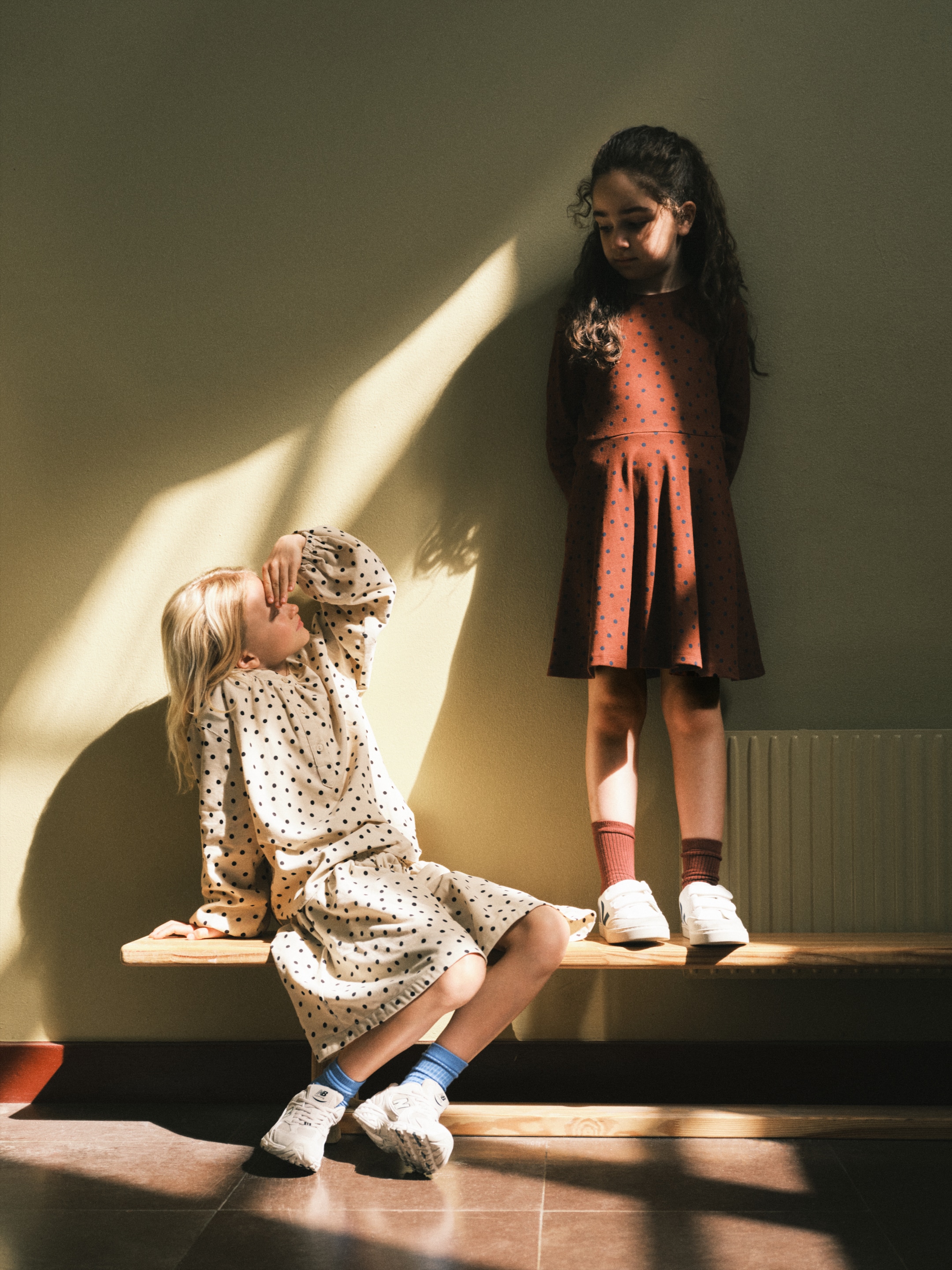 Two children in polka dot dresses stand and sit on a wooden bench in soft, dappled sunlight, creating a calm, serene atmosphere indoors.