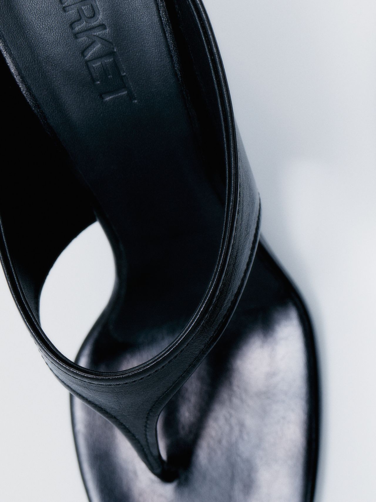 Close-up of a black leather sandal with a sleek strap and subtle shine, showing a part of the footbed with embossed text.