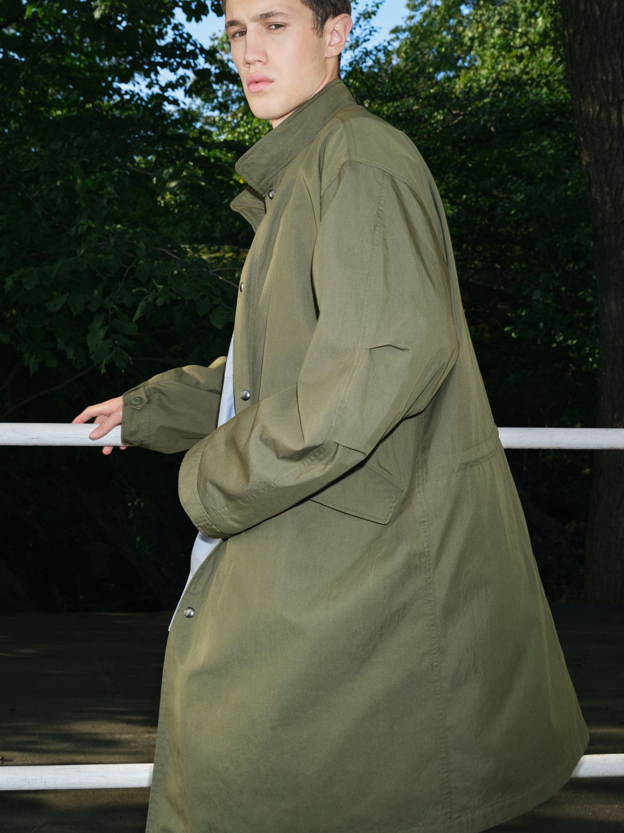 Person in a long olive-green coat stands outdoors, holding a railing, with lush green trees in the background.