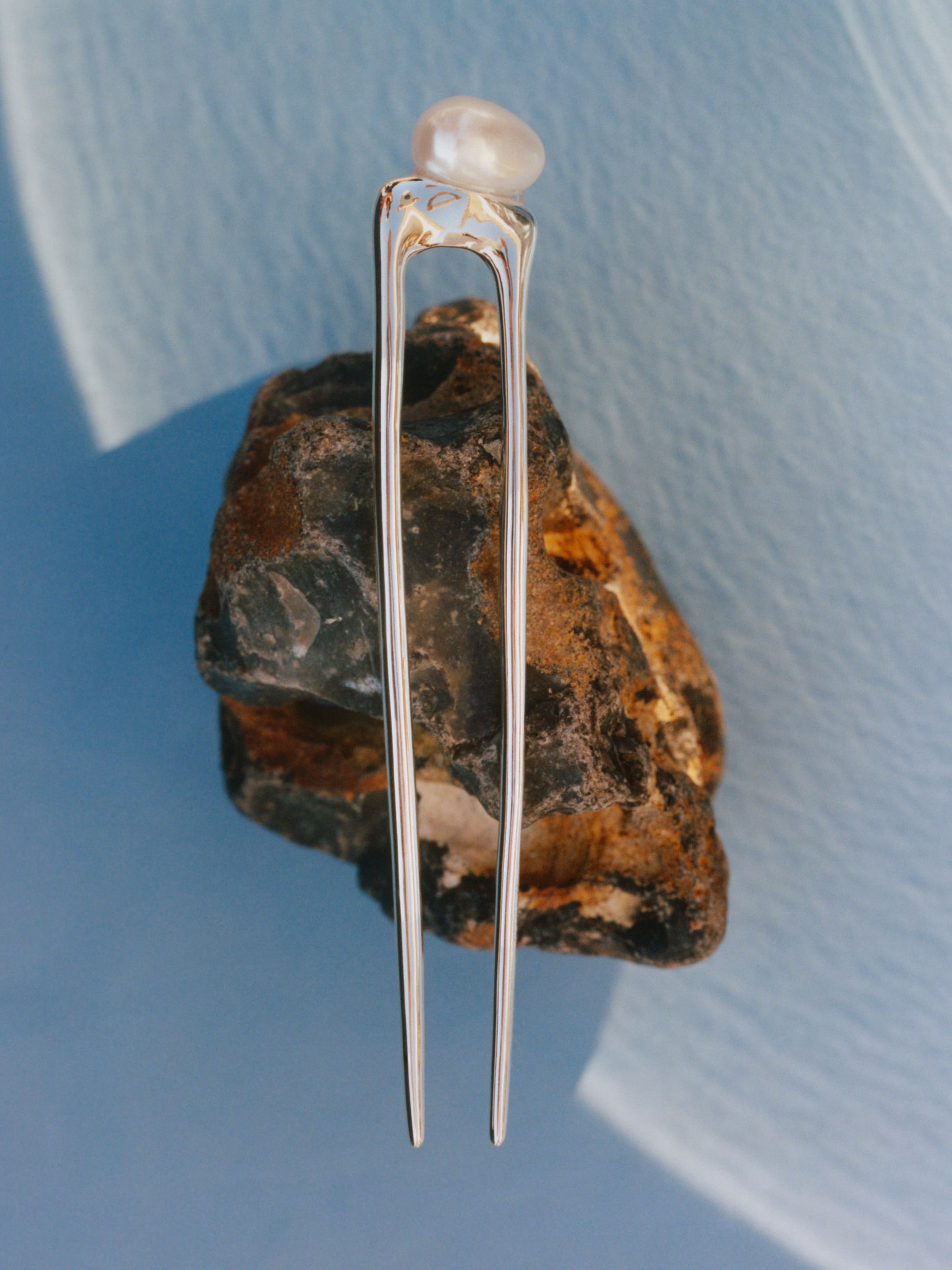 A silver hairpin with an elongated design and a pearl on top, resting on a textured rock against a light blue background.