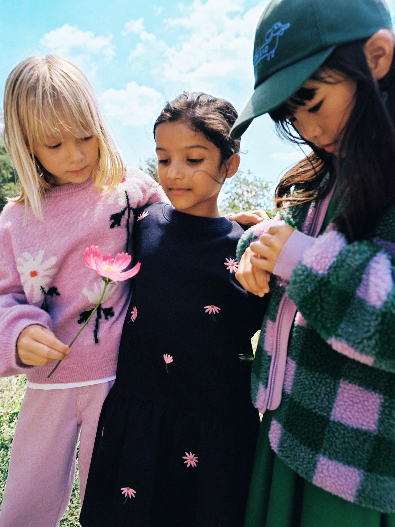Three children outdoors, one holding a pink flower. They wear colorful clothing, including a sweater, dress, and cap. Blue sky in the background.