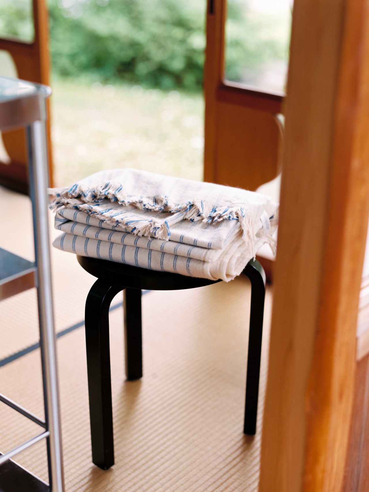 A stack of folded, fringed textiles with blue stripes sits on a black stool in a sunlit room with a view of greenery outside.