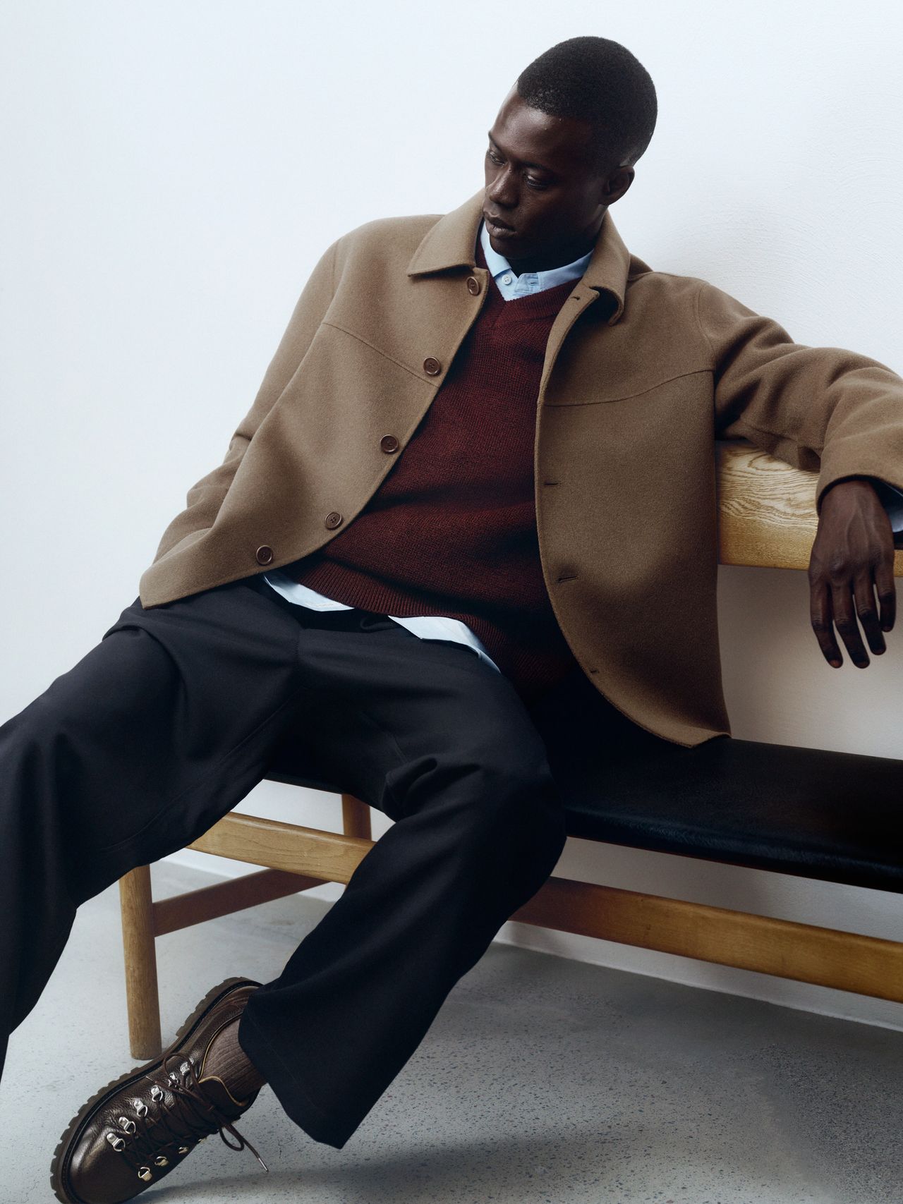 Man in a brown coat and dark pants sits on a wooden bench against a white wall, wearing a maroon sweater and brown shoes.