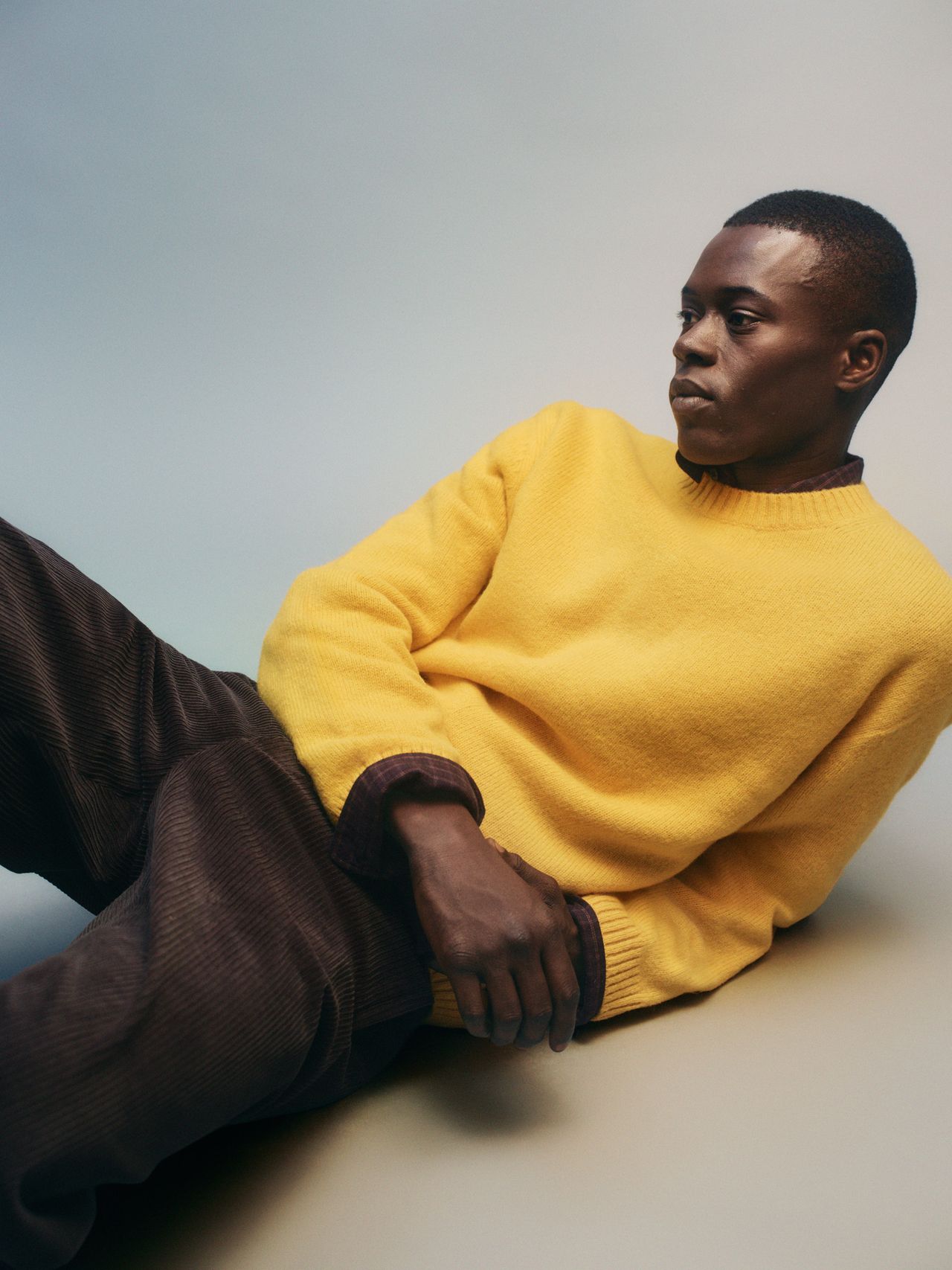 Person in a yellow sweater and brown pants, reclining on the floor against a neutral background, looking to the side.