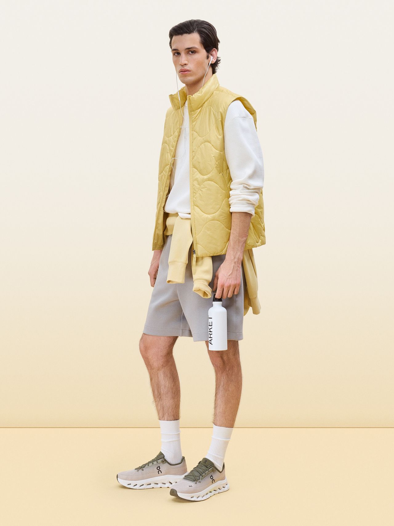 Young man in a yellow vest over a white shirt and gray shorts stands holding a white water bottle. The background is a soft, pale gradient.