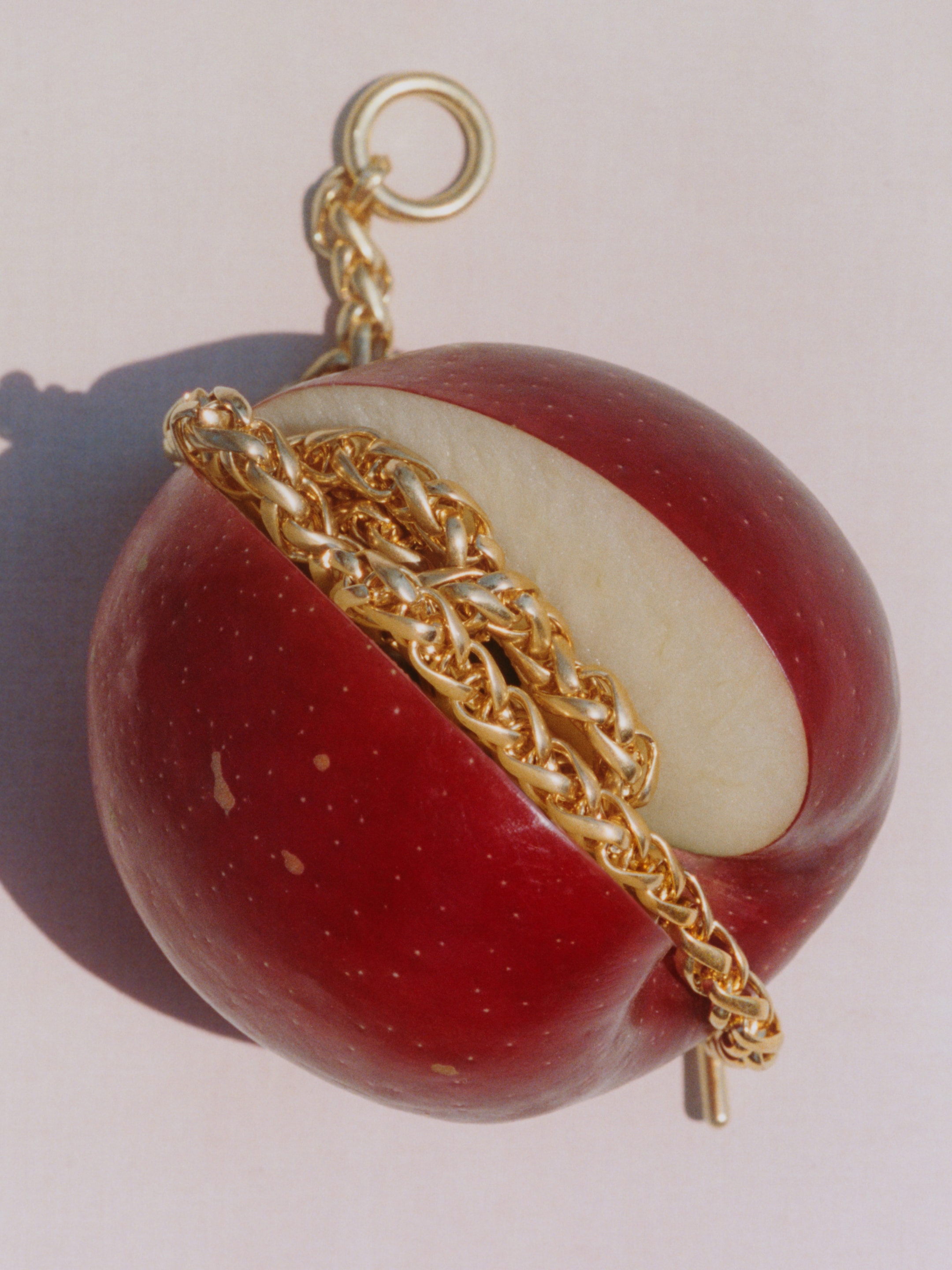 A red apple sliced open, with a gold chain intricately wrapped around it against a neutral background.