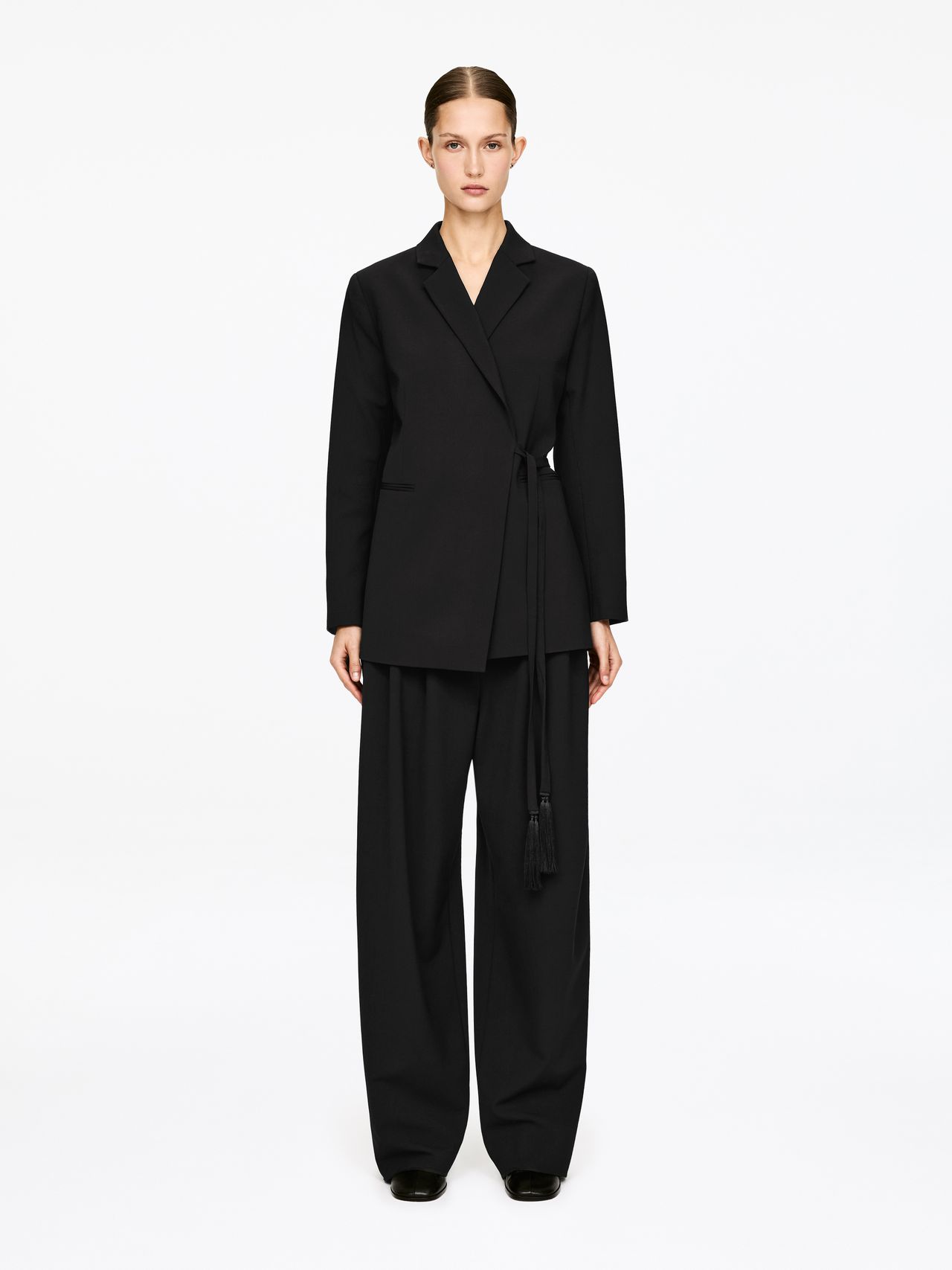 Model wearing a black tailored wrap blazer with tassel belt and wide-leg trousers, hair pulled back, on a plain white background.