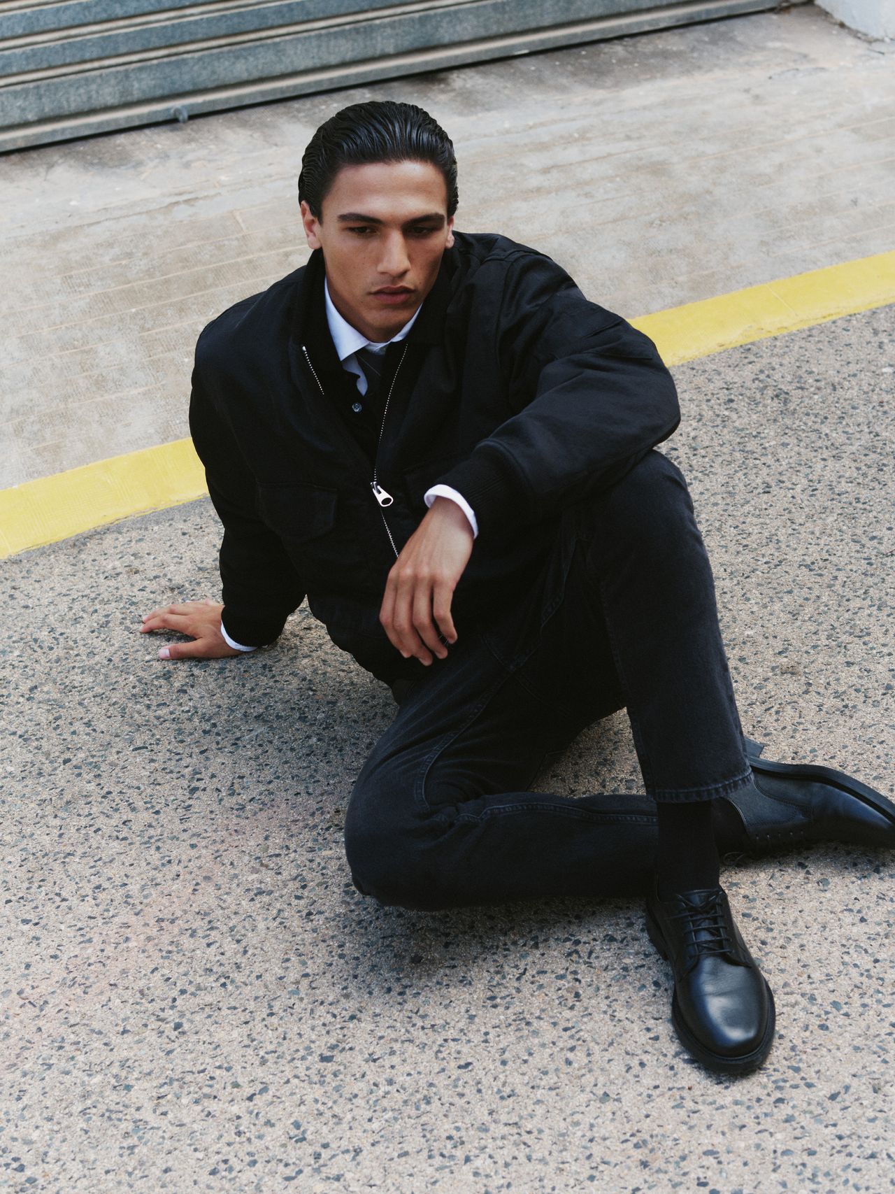 Person in black outfit sitting on pavement, leaning back on one arm with legs bent, near a yellow line.