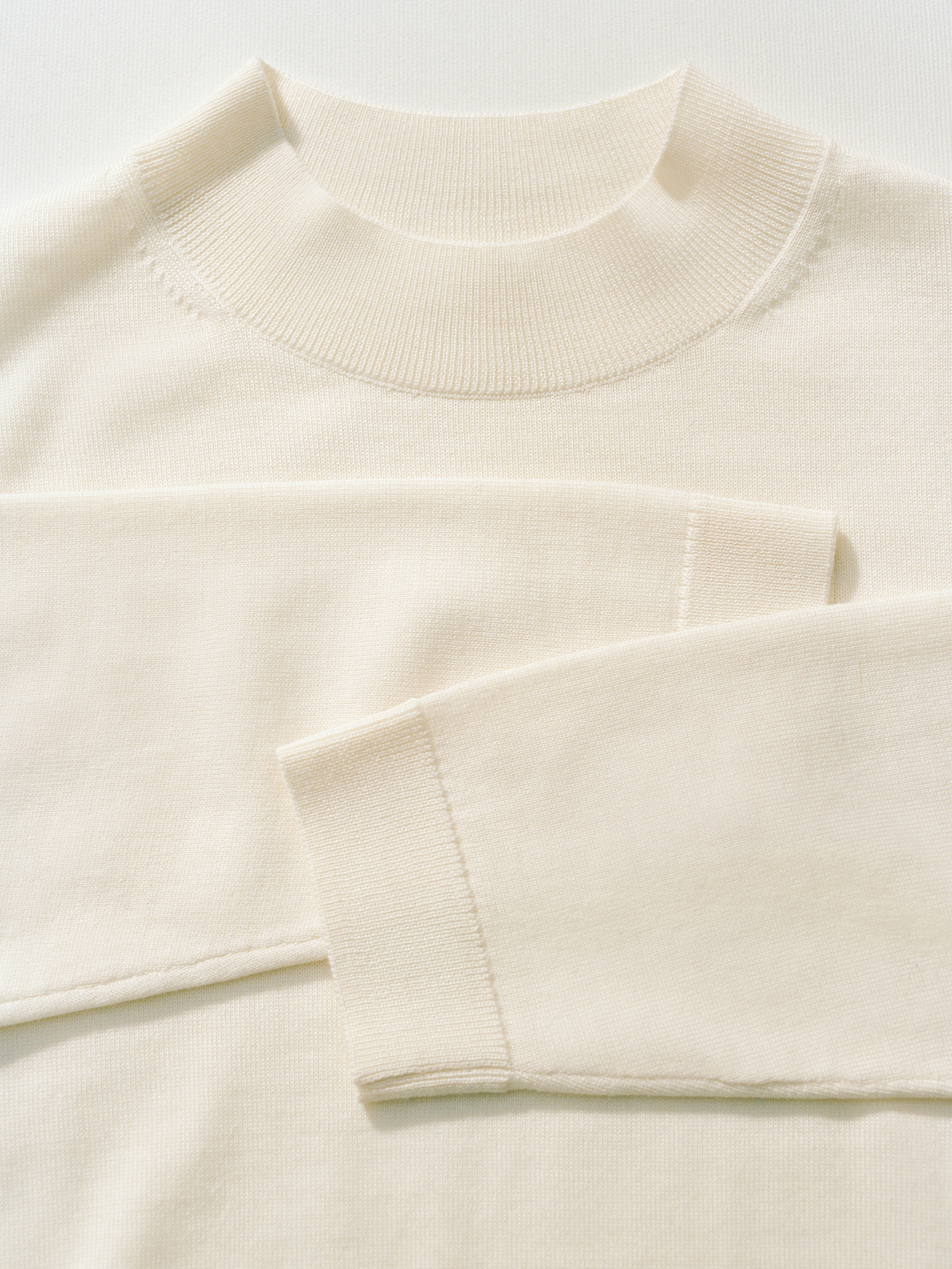 Close-up of a cream-colored, long-sleeve knit sweater, showing its ribbed collar and cuff details on a light background.