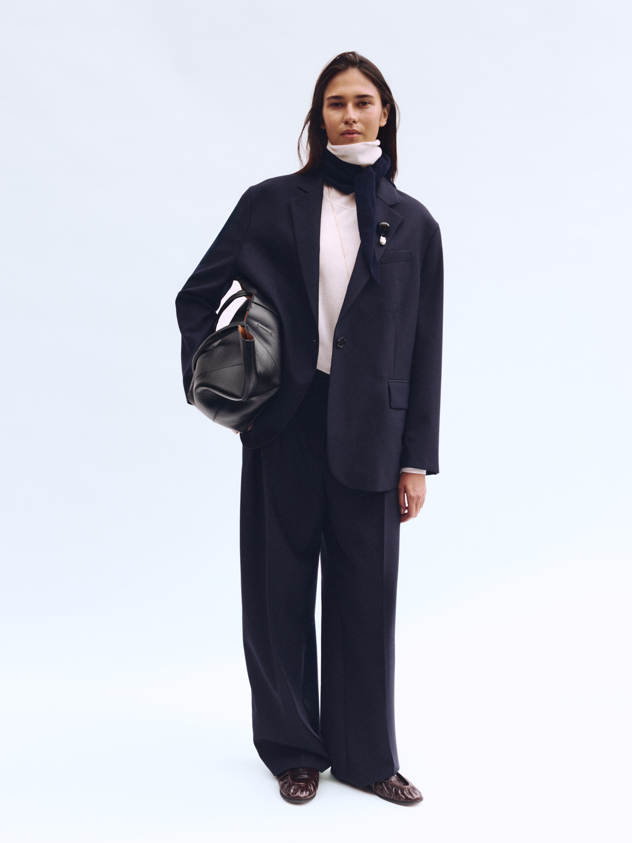 A person stands confidently wearing a dark suit, white shirt, and scarf, holding a black bag, against a plain light background.
