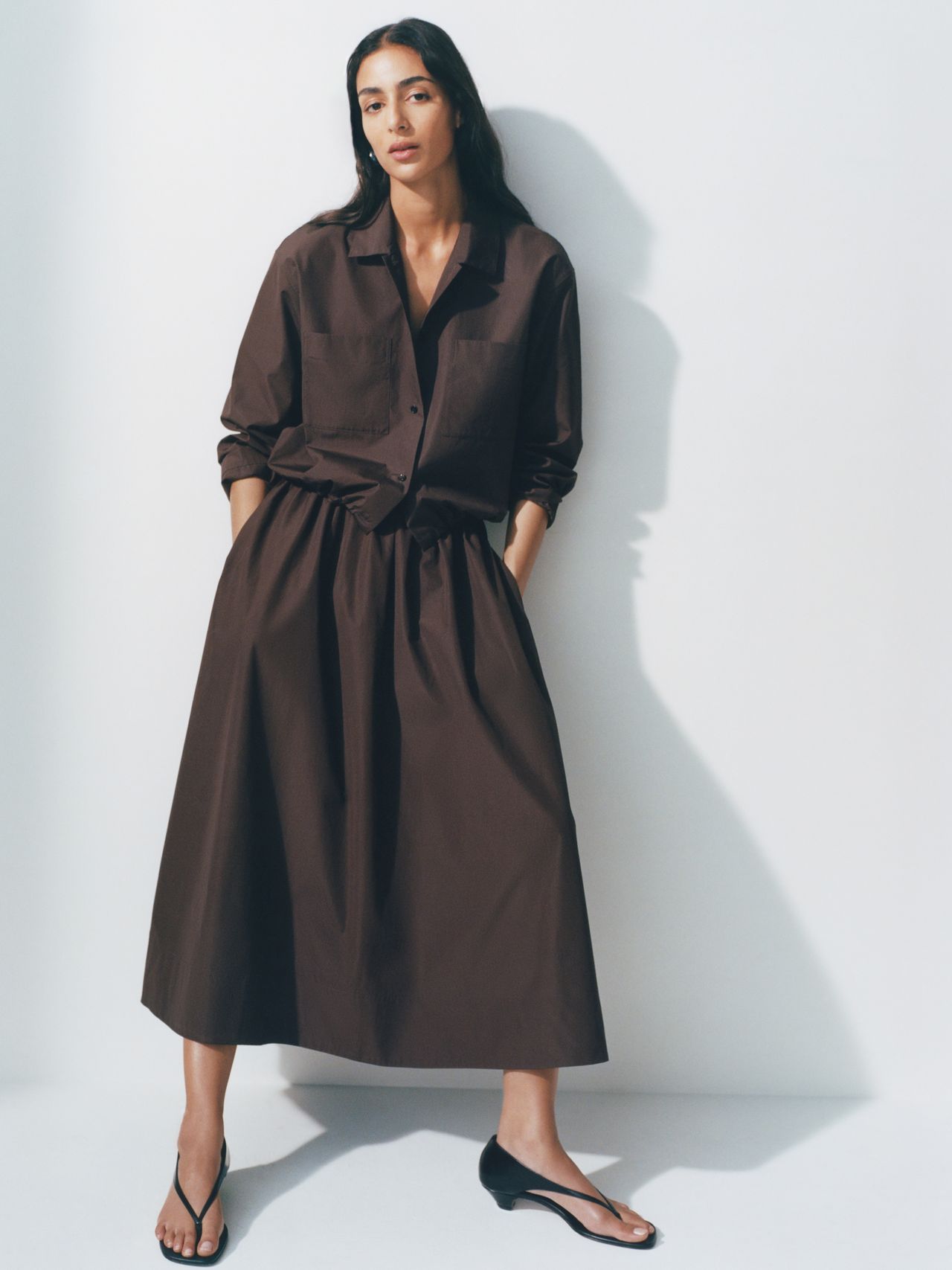 Woman in a long brown dress and black sandals, standing against a white background with hands in pockets, looking at the camera.